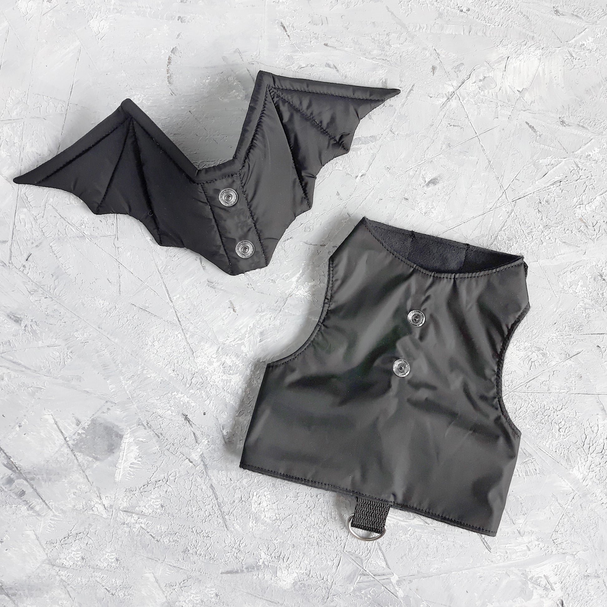 Overhead view showing the separate components of a black cat harness with bat wings. One piece is the vest-style harness body with snap closures and a D-ring, and the other is the bat wing attachment with corresponding snaps