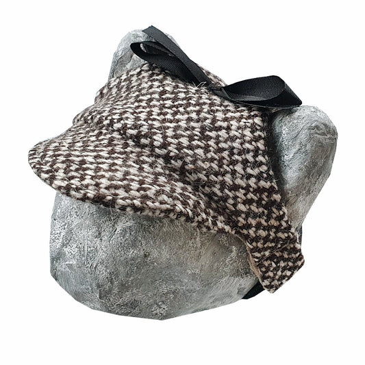 Tweed-style hat for a cat with a small brim and a black ribbon tied in a bow on top, shown on a grey textured surface.