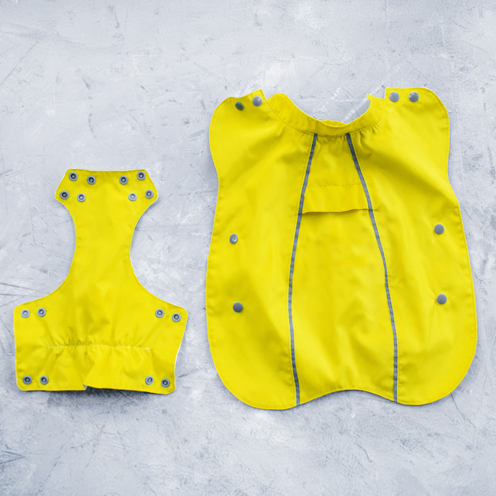 Flat lay of a bright yellow cat raincoat showing the main body and a separate piece, with reflective stripes and grey snap closures, on a textured grey surface.