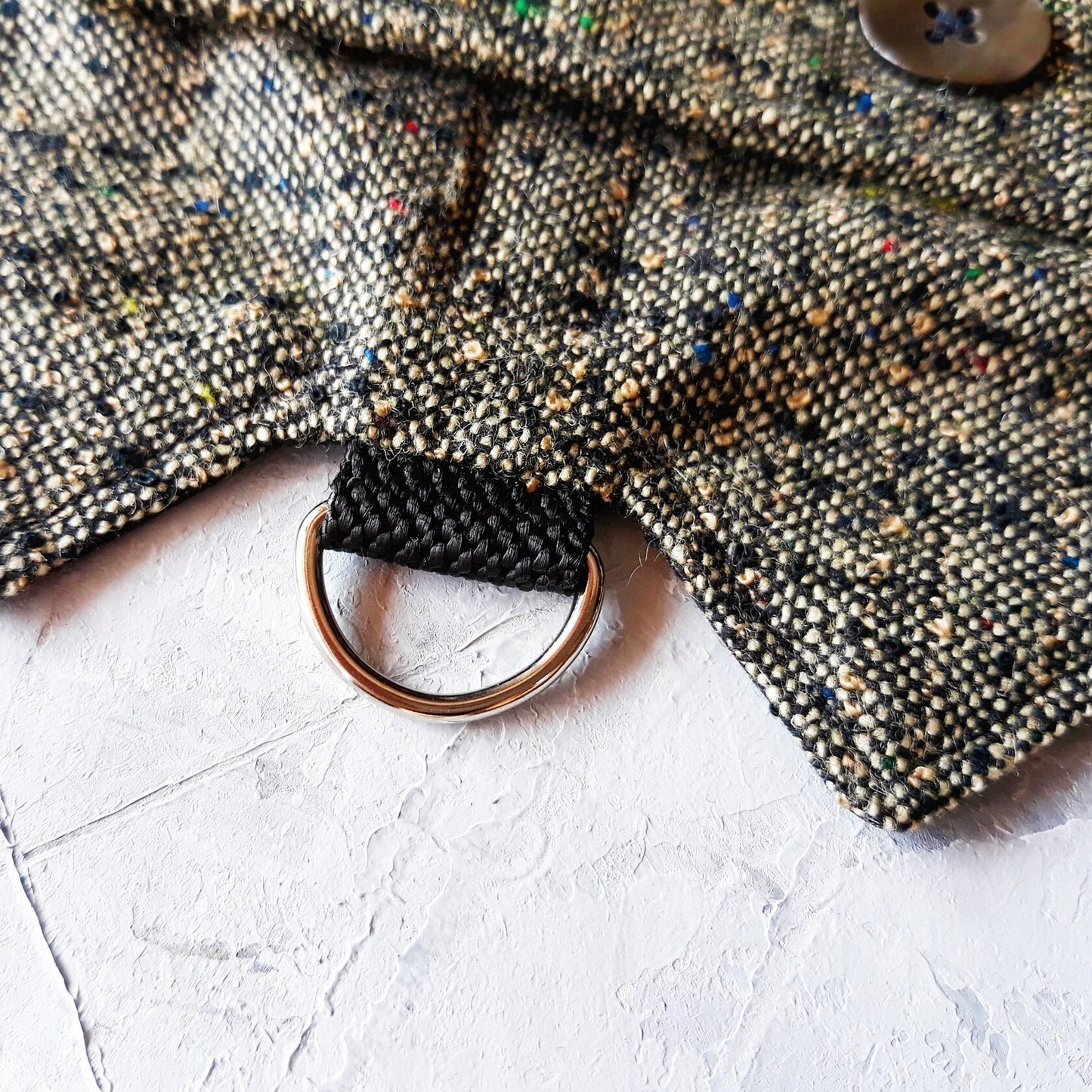 Close-up of a silver metal D-ring attached to a black strap on a grey tweed cat harness with colorful flecks.