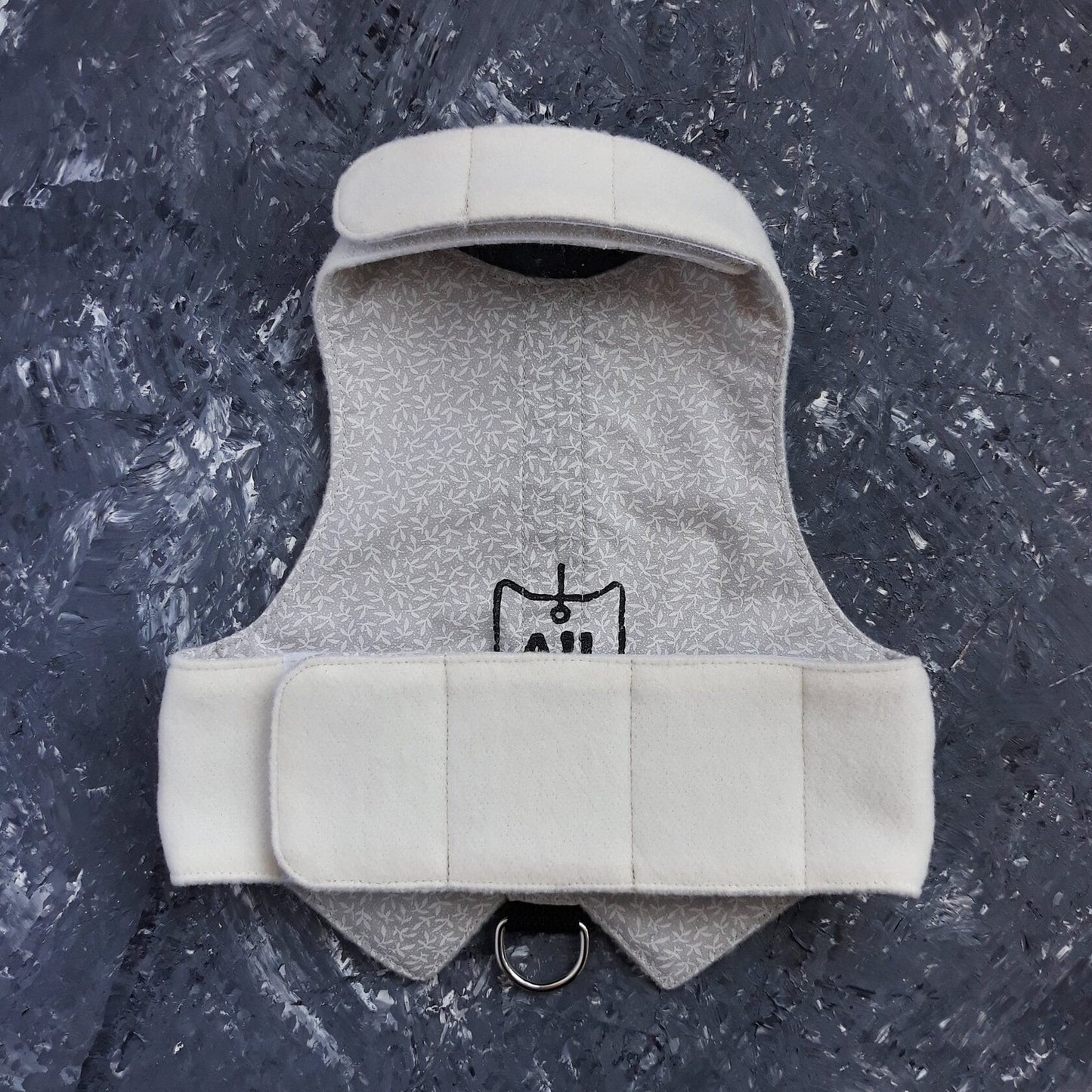 An overhead view of the inner lining of a white cat harness. The lining has a light grey pattern and features black text and a graphic. White Velcro straps and a metal D-ring are also visible.