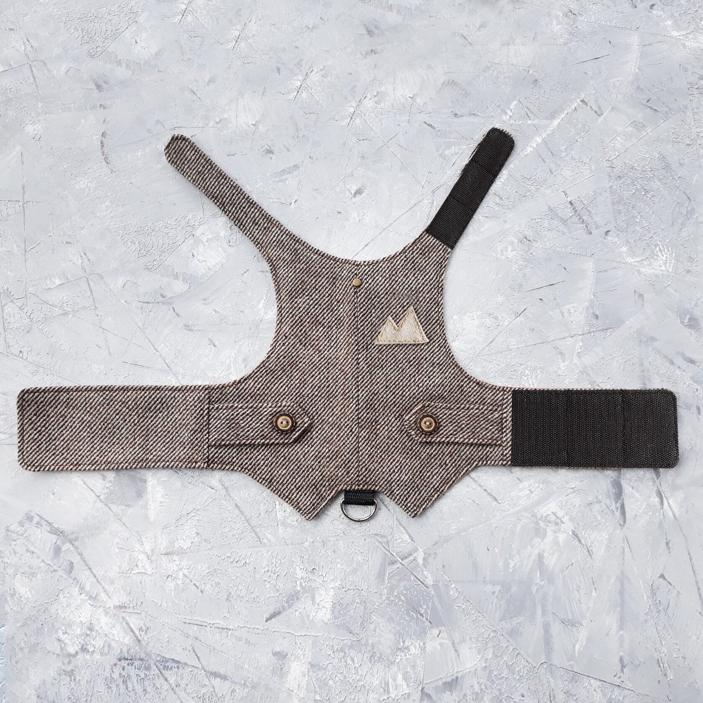 Overhead view of a brown tweed cat harness with a diagonal stripes pattern laid flat on a textured grey surface. The harness features decorative buttoned tabs, a light brown triangular patch, and adjustable brown Velcro straps. A black leash attachment point is visible.