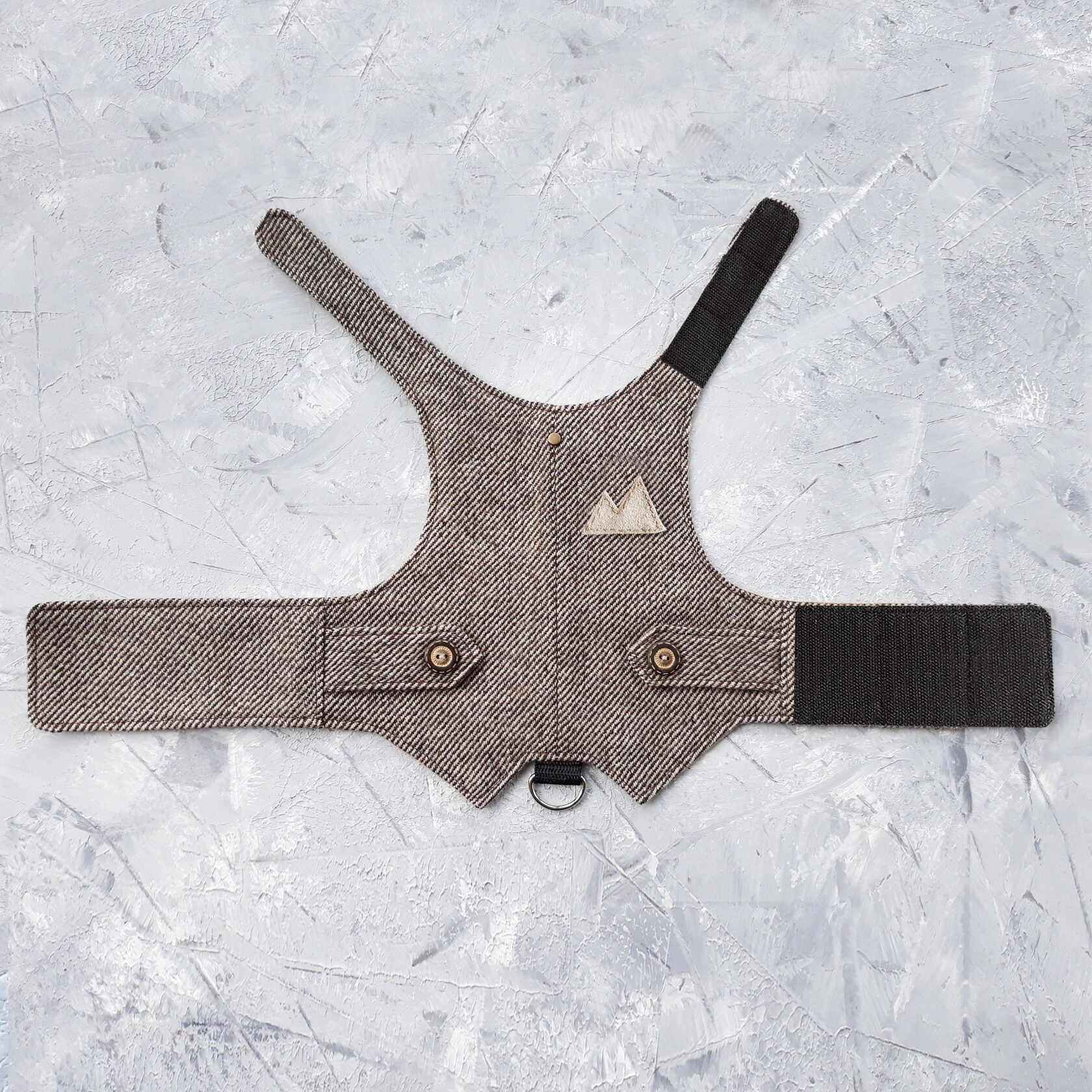 Overhead view of a brown tweed cat harness with a diagonal stripes pattern laid flat on a textured grey surface. The harness features decorative buttoned tabs, a light brown triangular patch, and adjustable brown Velcro straps. A black leash attachment point is visible.