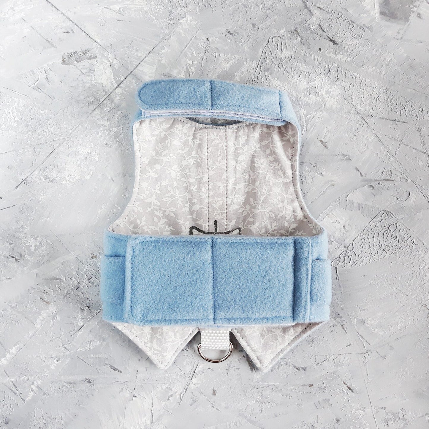 The inside of a light blue cat harness made of wool or felt, revealing a light grey lining with a subtle floral pattern.