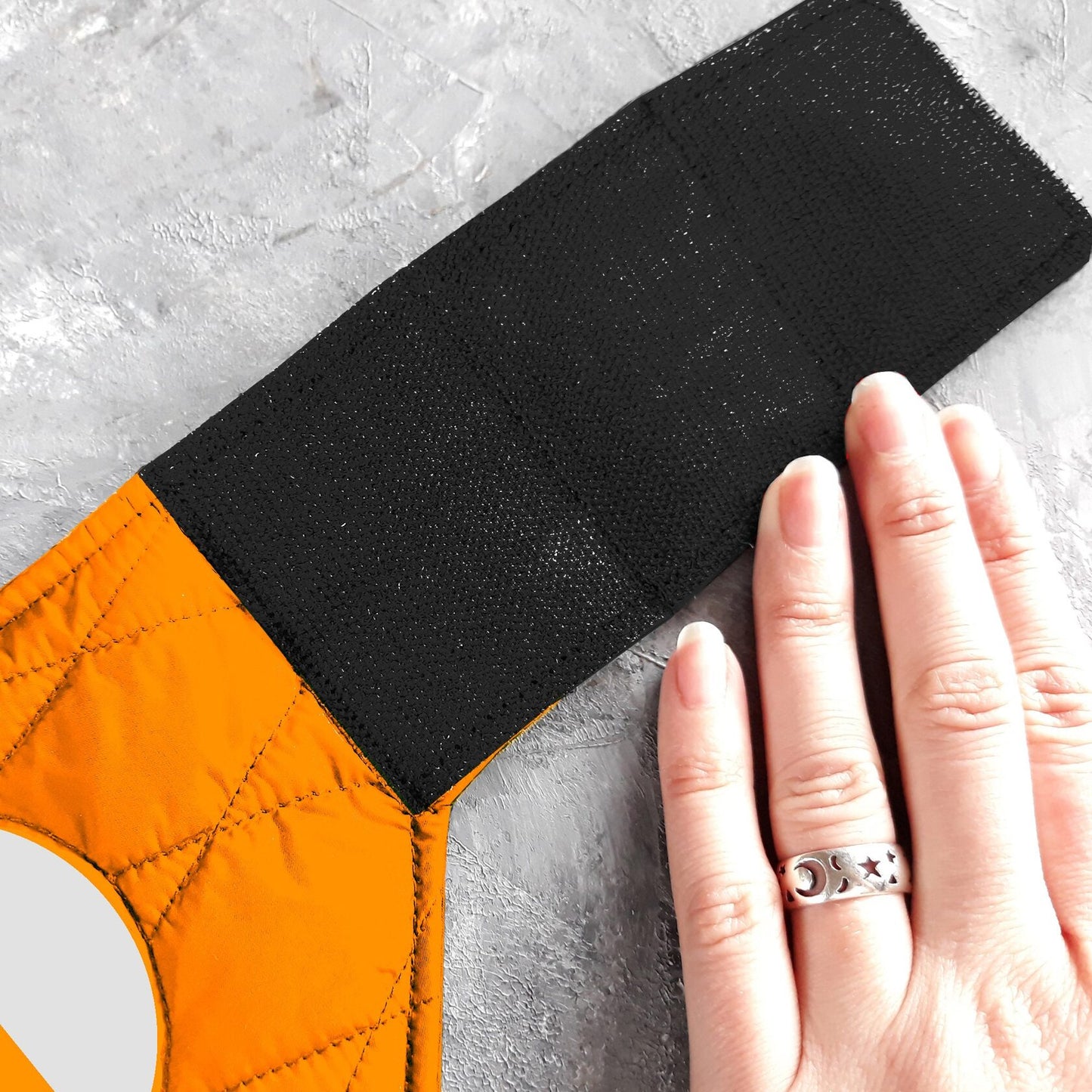 Close-up of a hand pressing down on the black Velcro closure of a quilted orange cat harness with a reflective element visible.