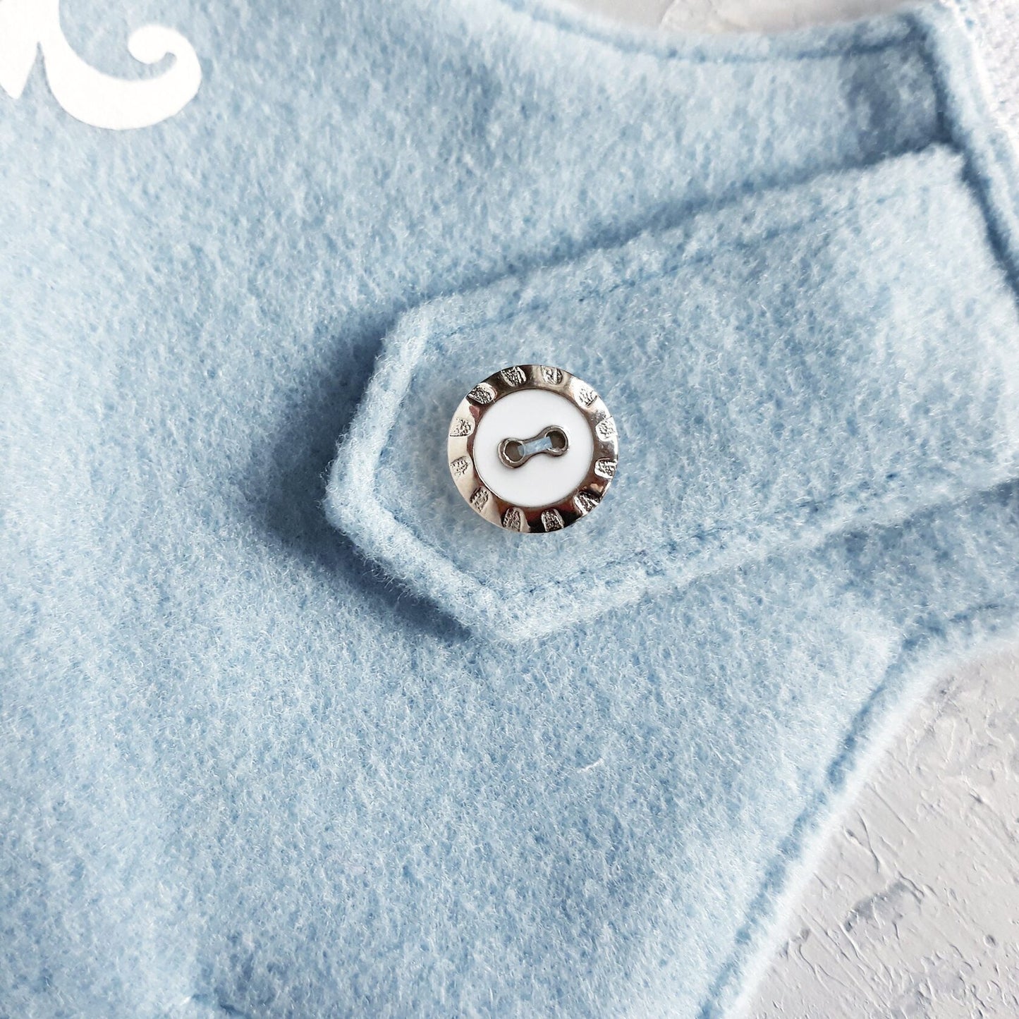 Silver-rimmed white button with a paw print design, attached to a light blue cat harness flap.
