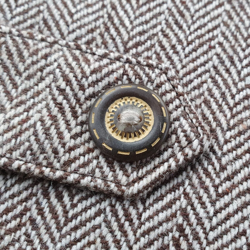 Close-up of a decorative bronze button with a textured center on a brown and cream herringbone tweed cat harness.