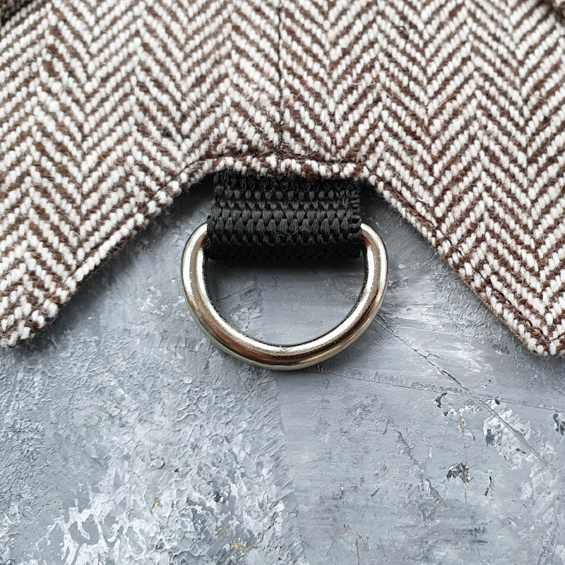 Close-up of a silver metal D-ring attached to a black strap on a brown and cream herringbone tweed cat harness.