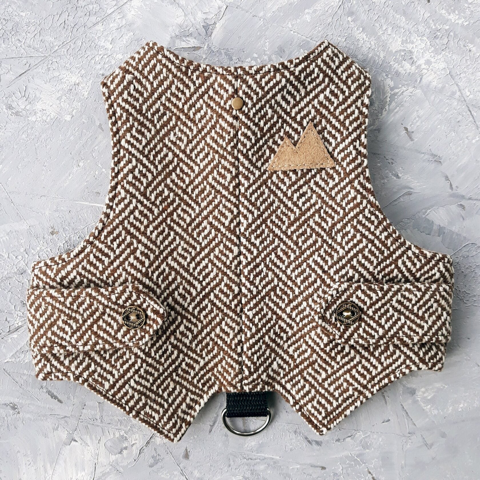 A cat harness made from brown tweed fabric with a distinctive geometric pattern. A functional D-ring connected by a black strap.
