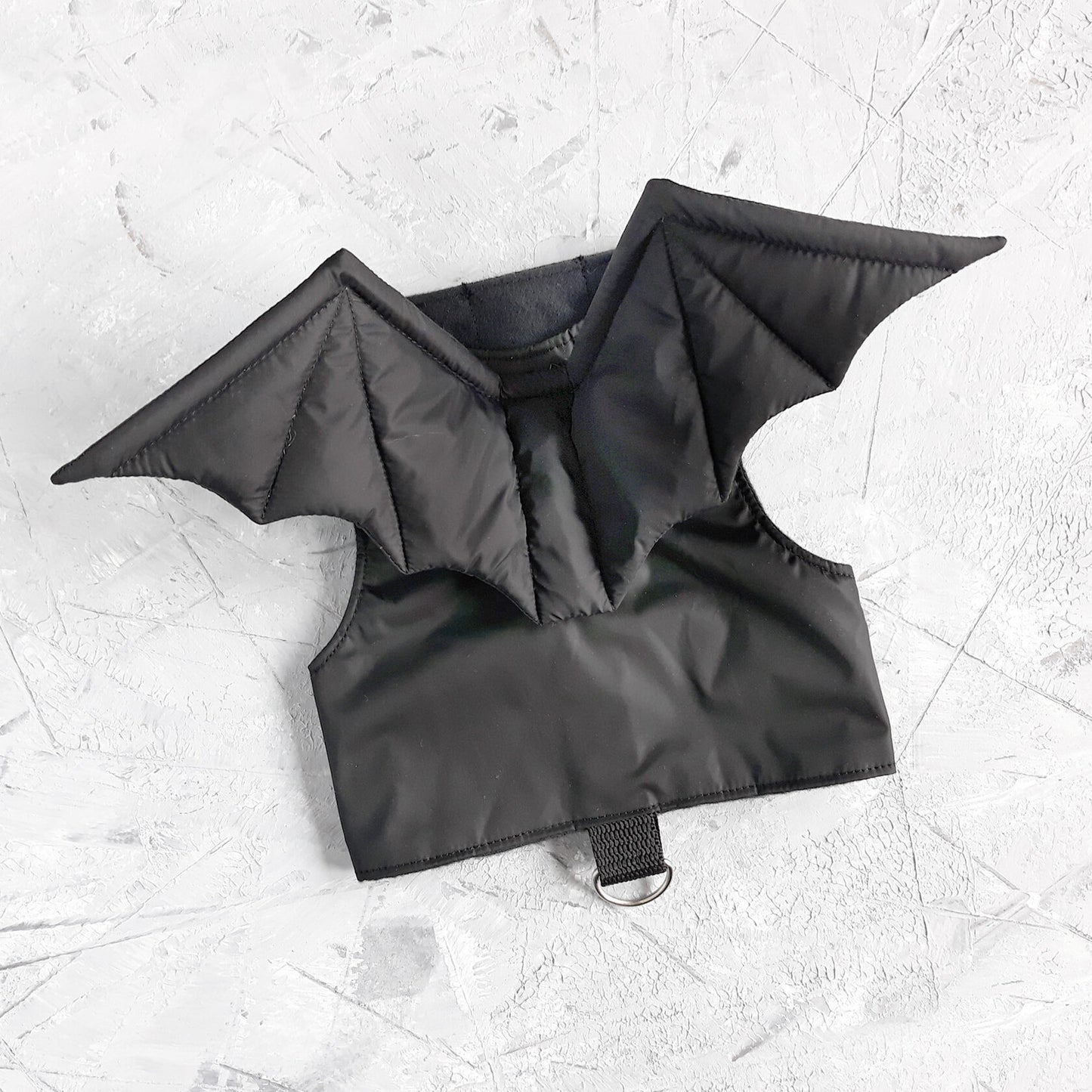 A black cat harness with a distinct design that includes two bat wings extending from the back area. The harness has straps that would secure around the cat's neck and chest, fastened with Velcro.