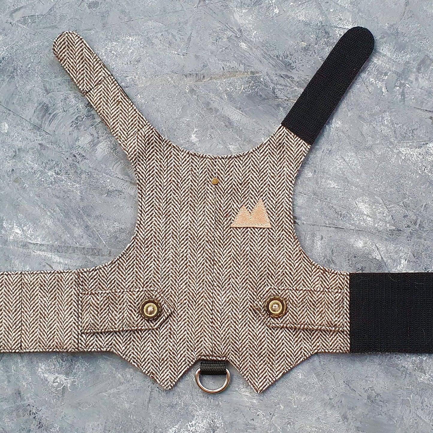 A brown and cream herringbone tweed cat harness with decorative bronze buttons, a brown faux leather mountain-shaped patch, and a black D-ring.