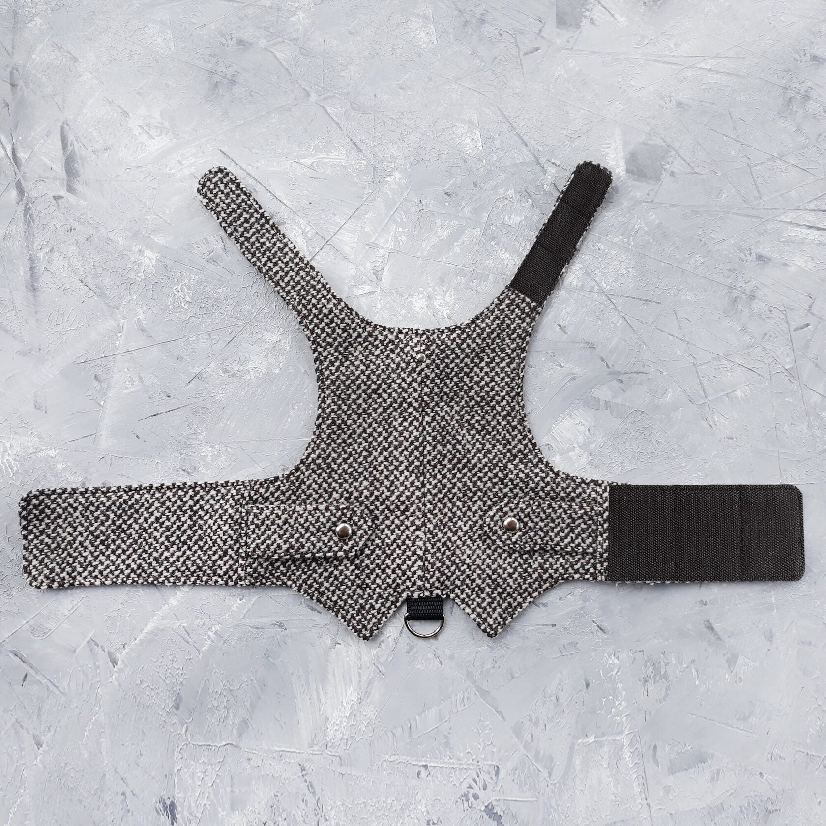 A flat lay of a black and white houndstooth cat harness with silver decorative buttons, black Velcro straps, and a D-ring.