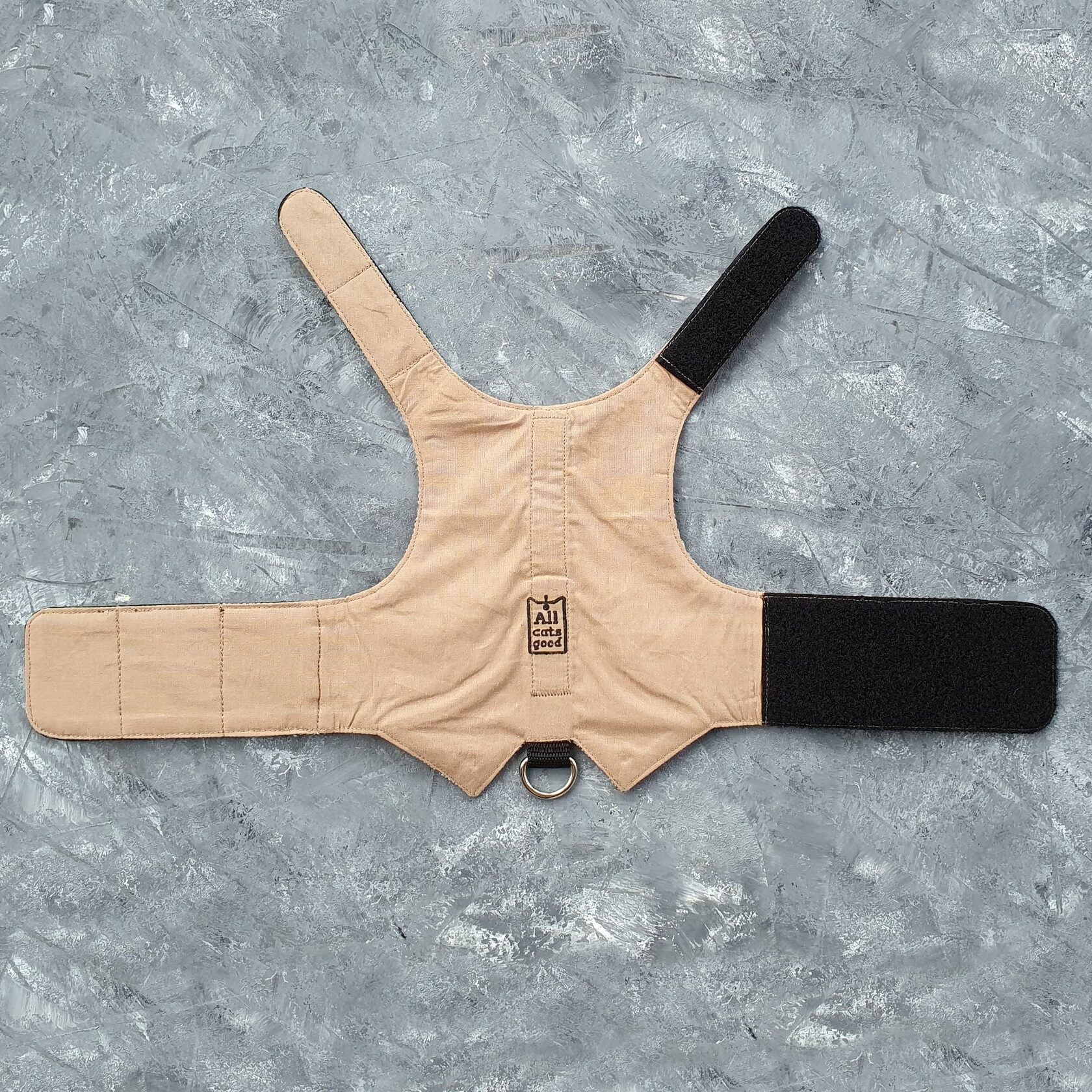 The inside of a brown herringbone tweed cat harness showing a plain beige fabric lining, black Velcro straps, and a black D-ring attachment. A small "Allcatsgood" tag is visible.