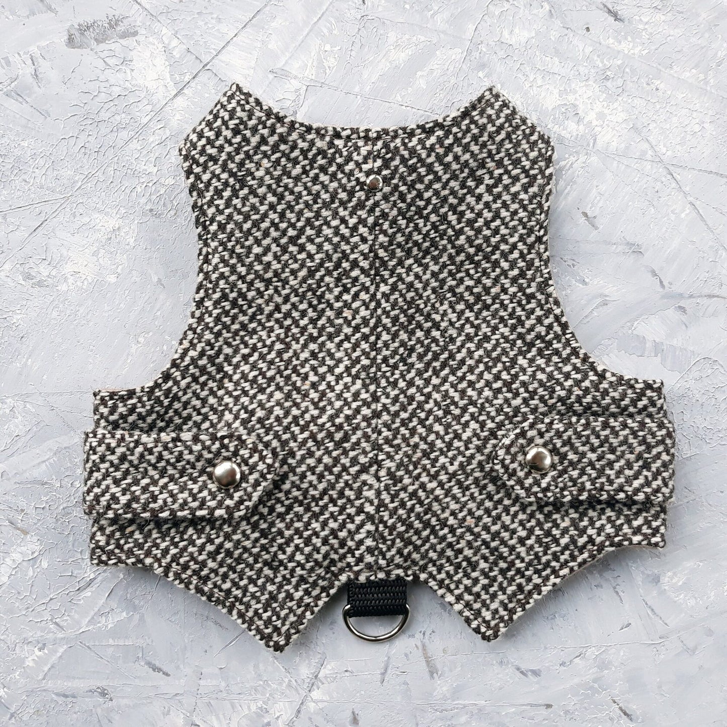 A black and white houndstooth cat harness with silver decorative buttons and a black D-ring, laid flat on a grey surface.