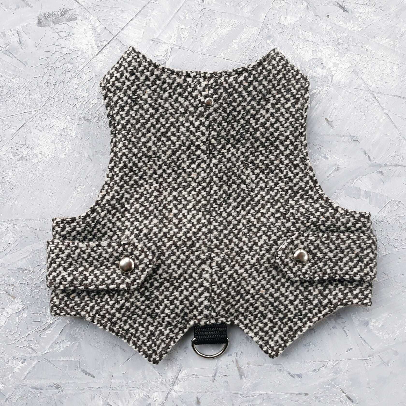 A black and white houndstooth cat harness with silver decorative buttons and a black D-ring, laid flat on a grey surface.