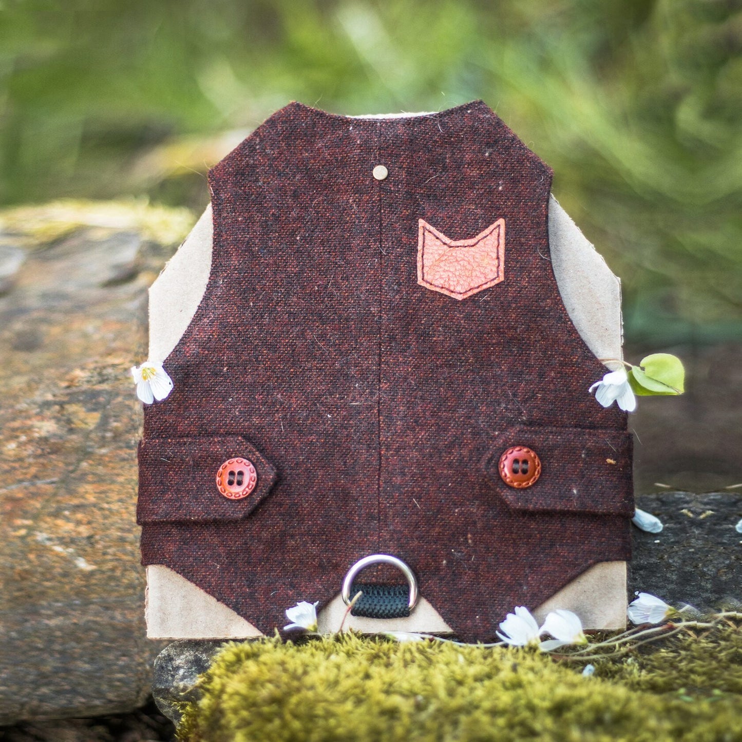 A burgundy tweed cat harness with a brown cat-shaped patch and decorative buttons, resting on moss-covered rocks with small white flowers.