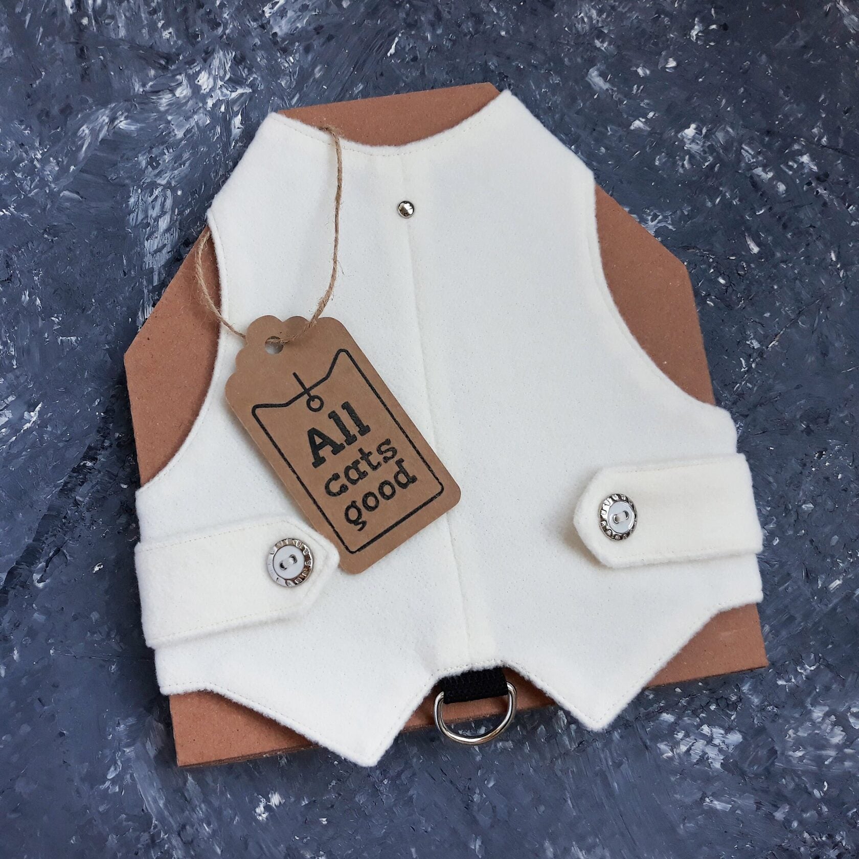 A white, classic-style cat harness from "Allcatsgood" is shown with a product tag bearing the brand's name. The harness has a clean design with snap closures for easy fastening and a D-ring for leash attachment.