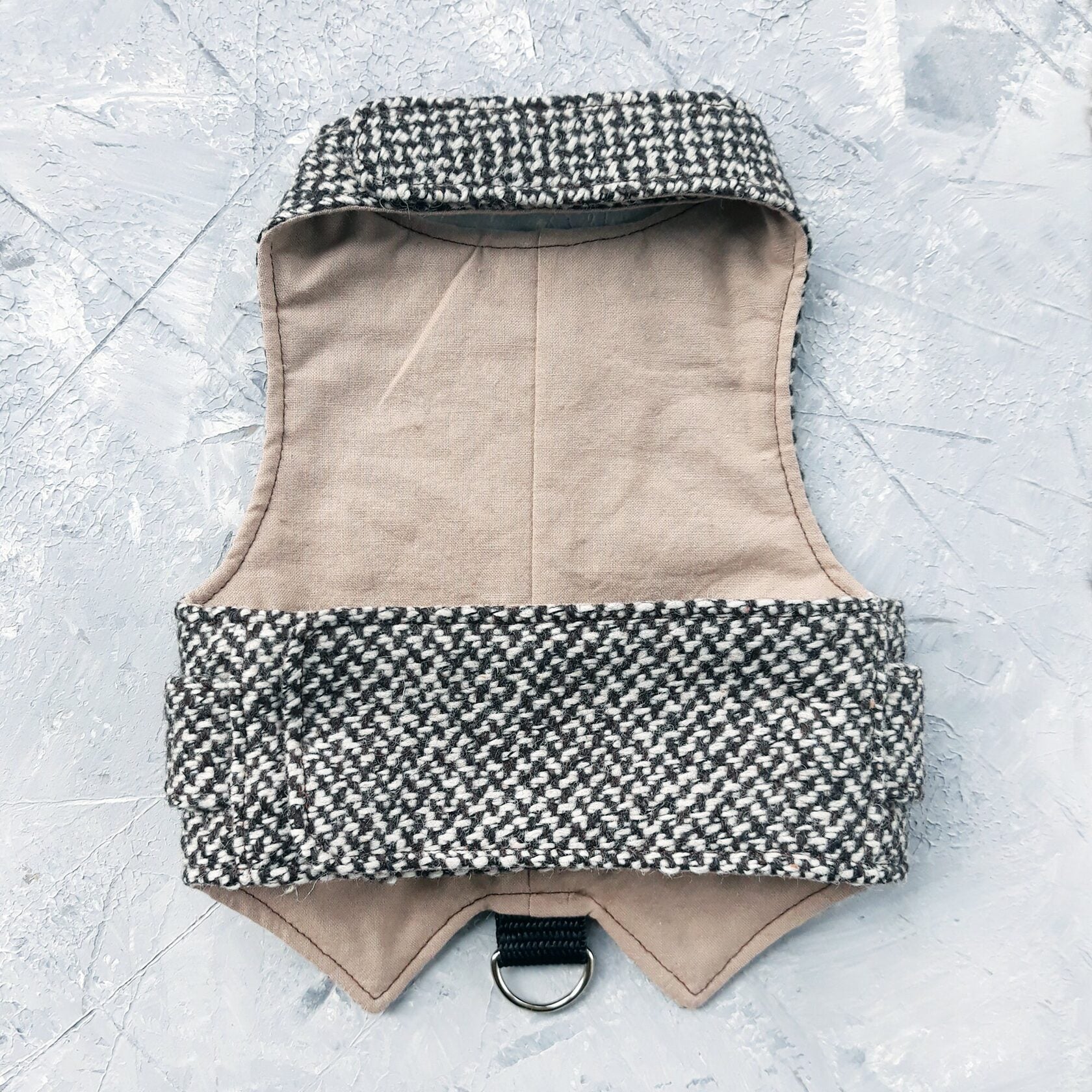 The inside of a black and white houndstooth cat harness showing a plain beige fabric lining and a black D-ring attachment.