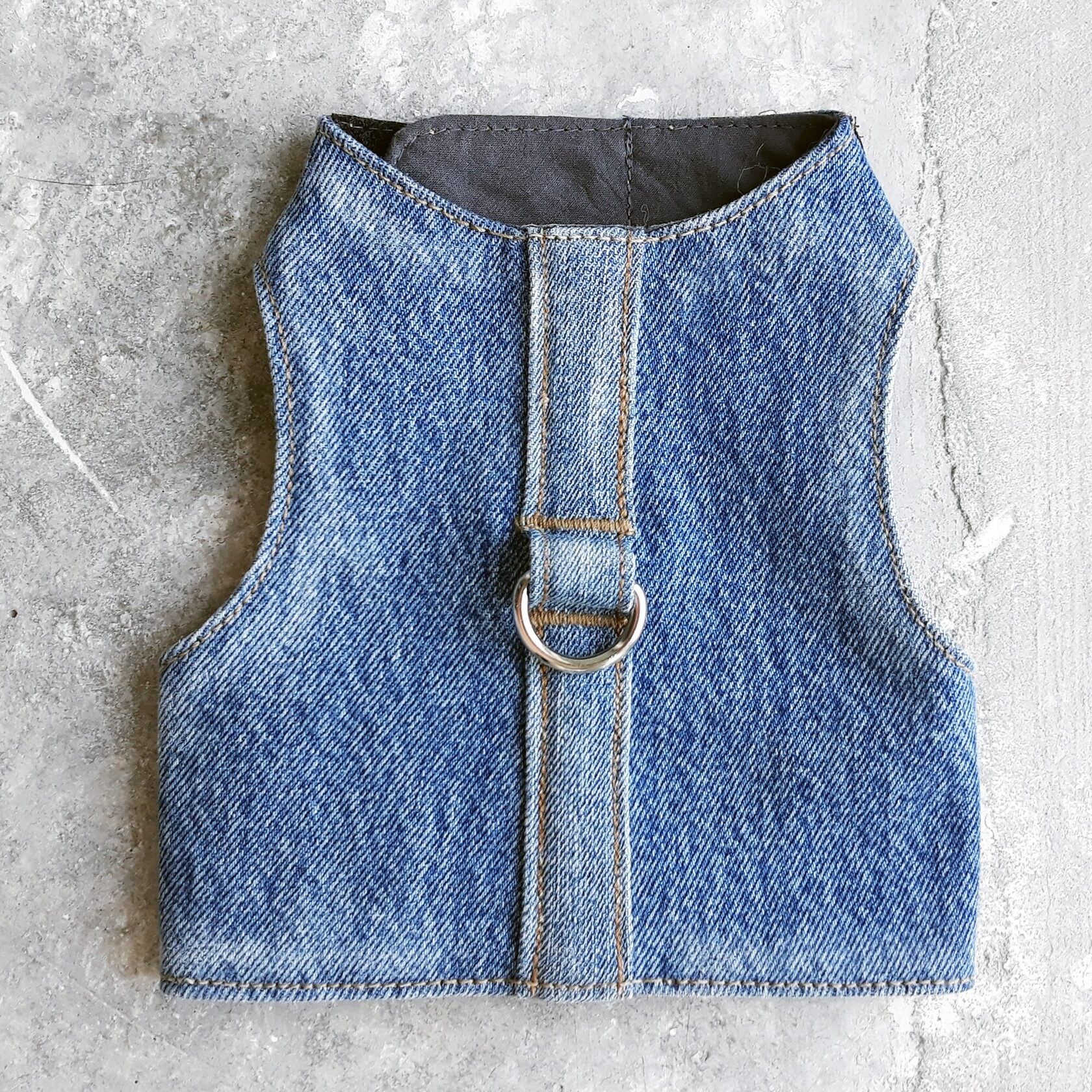 A cat harness made from blue denim fabric, featuring a central strap and a silver-colored D-ring.
