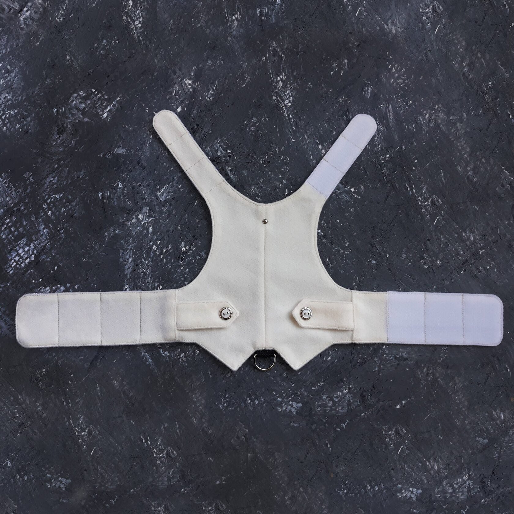 A white cat harness is laid out to display its vest shape, white Velcro straps for adjustability, snap closures for fastening, and a metal D-ring for attaching a leash.