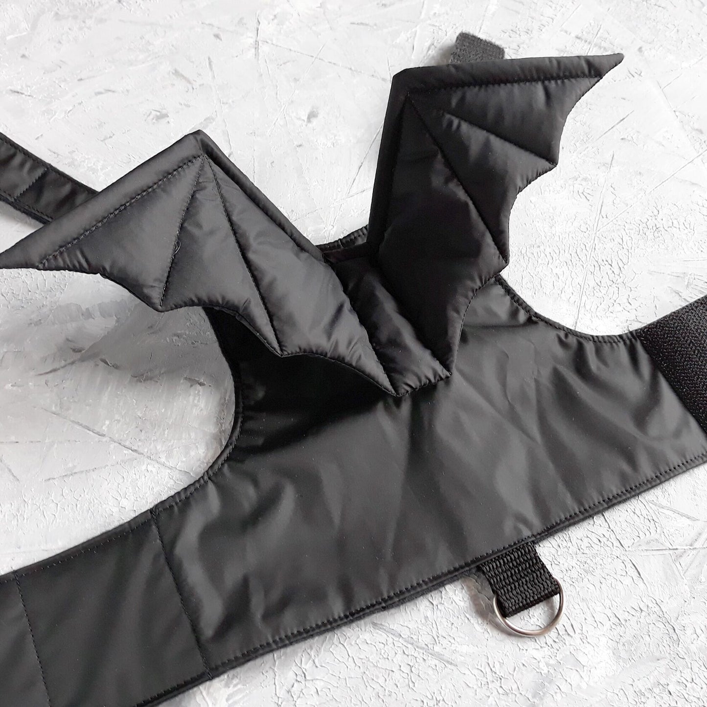 A black cat harness with a distinct design that includes two bat wings extending from the back area. The harness has straps that would secure around the cat's neck and chest, fastened with Velcro.