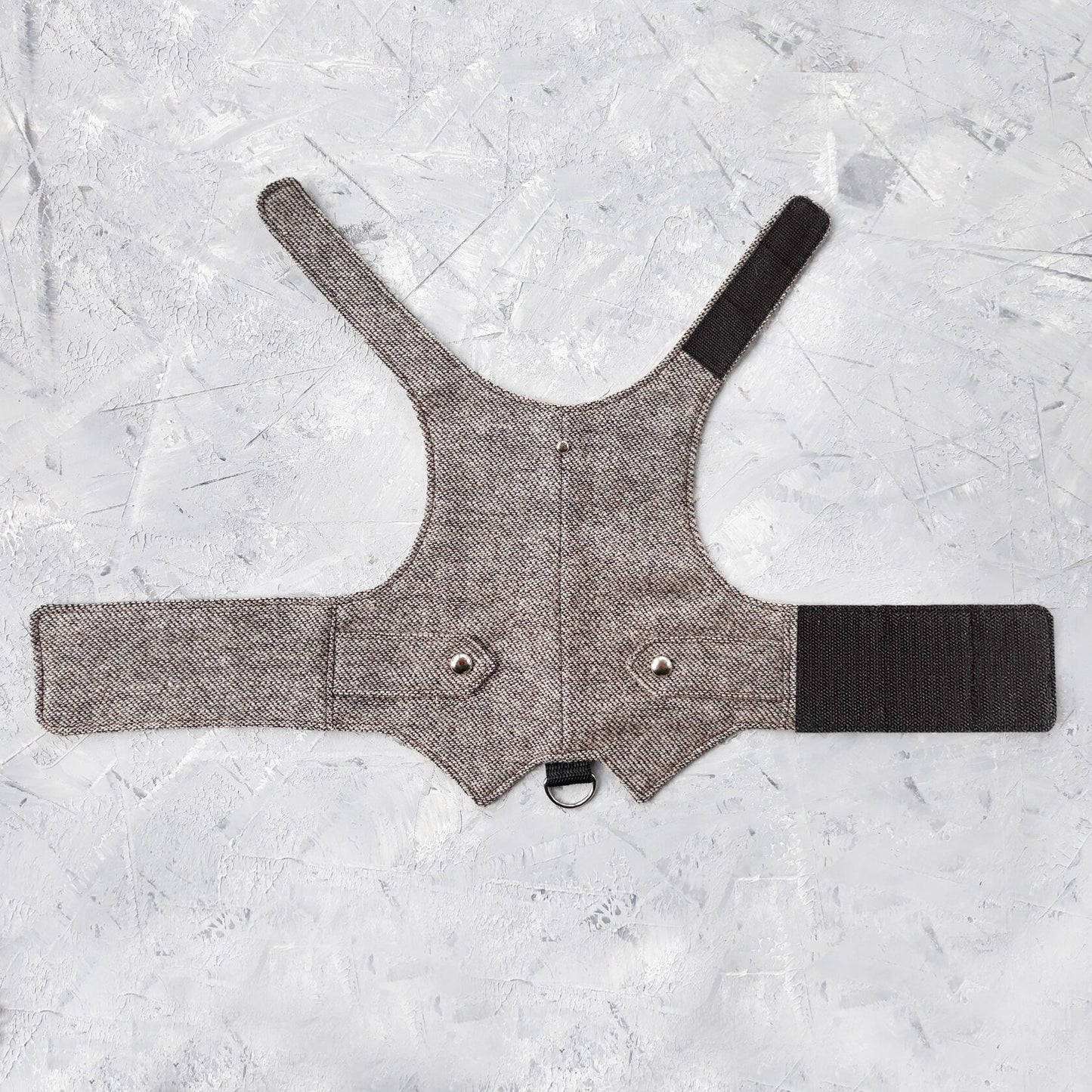 A flat lay of a light brown tweed cat harness with silver decorative buttons, black Velcro straps, and a D-ring.