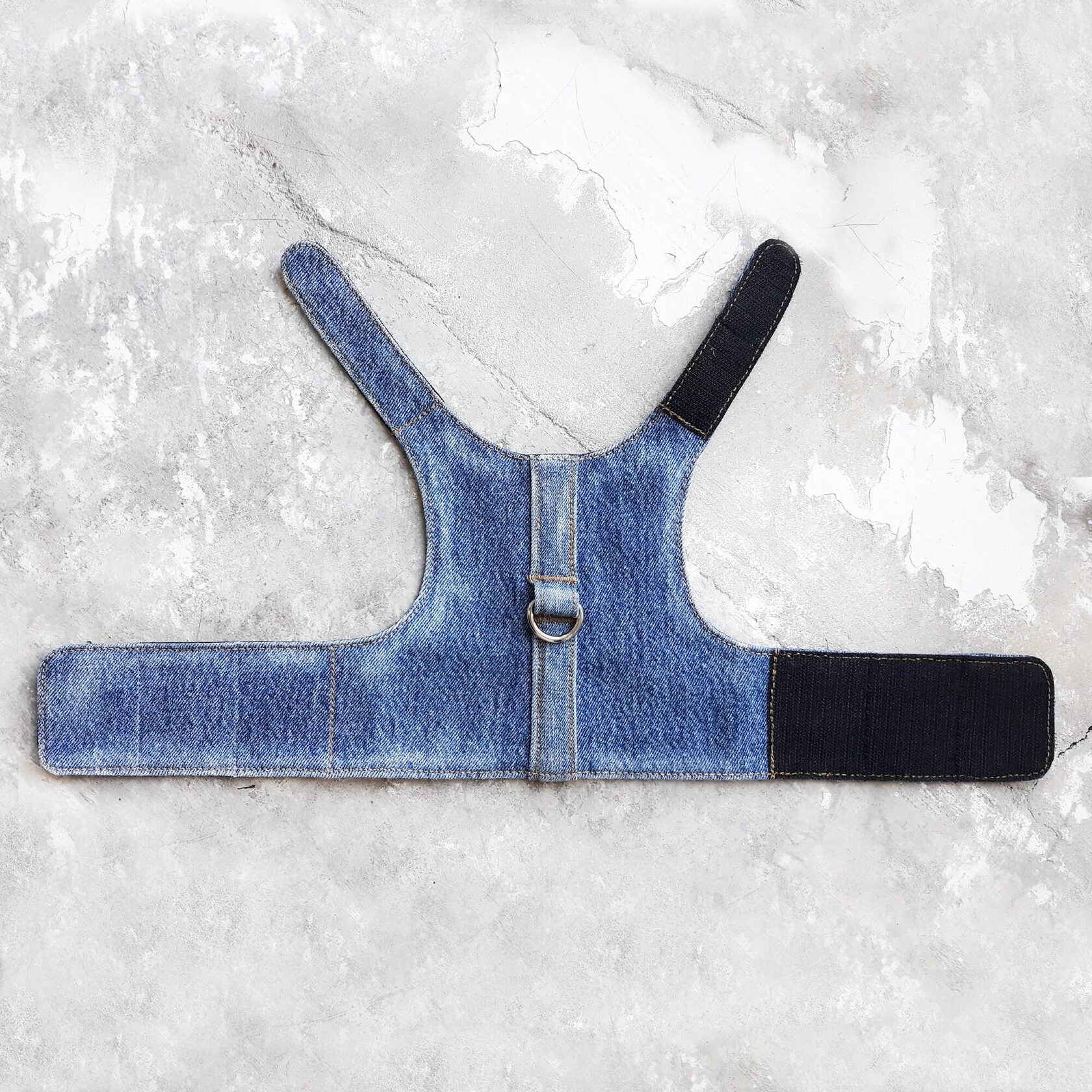 A blue denim vest-style harness designed for cats is shown flat, featuring a central vertical denim strap, a metal D-ring, and black Velcro straps for securing the harness around the cat's body.