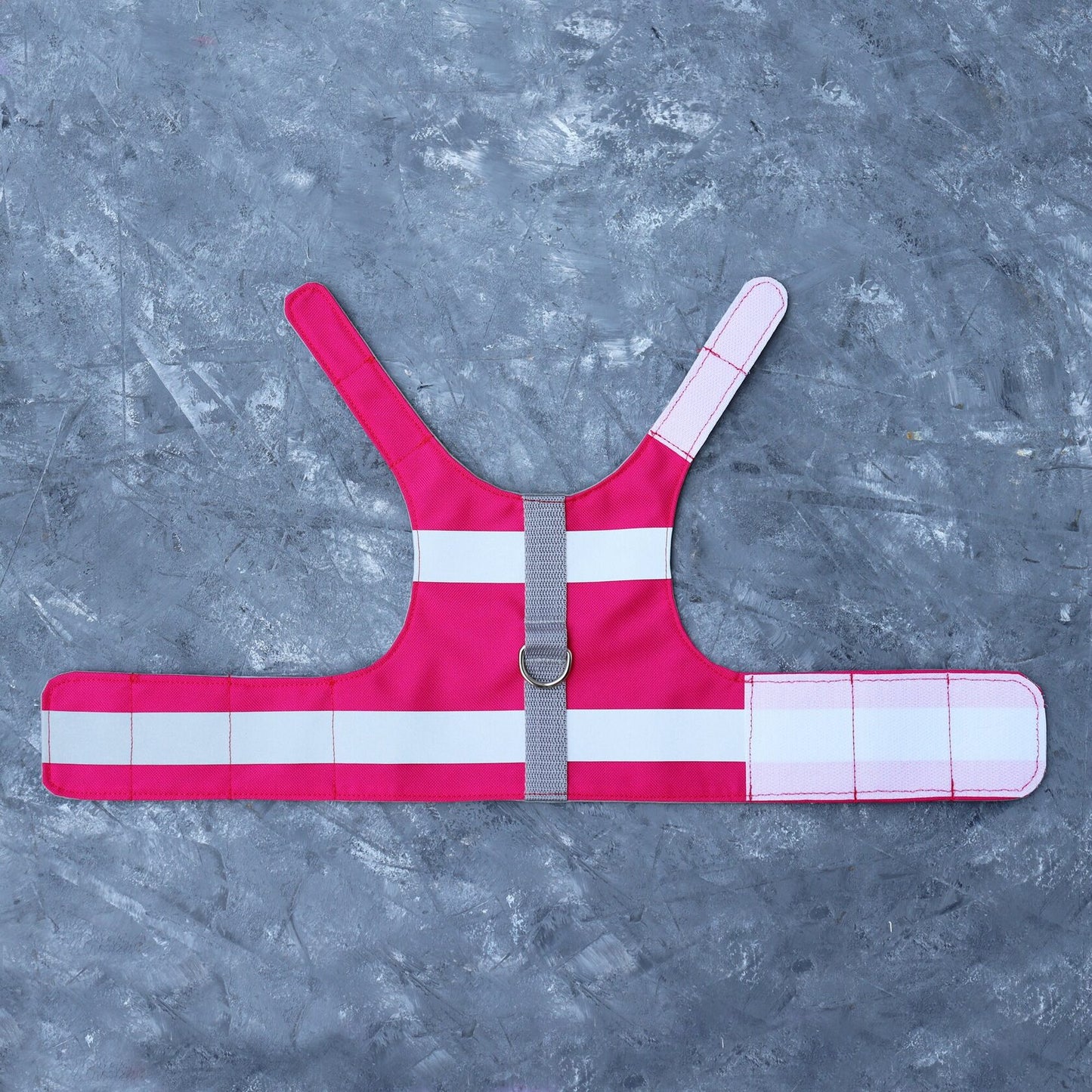 Overhead view of a bright pink cat safety harness laid flat on a textured grey surface. The harness features two horizontal silver reflective stripes, adjustable light pink Velcro straps, a grey central strap, and a metal D-ring for leash attachment.