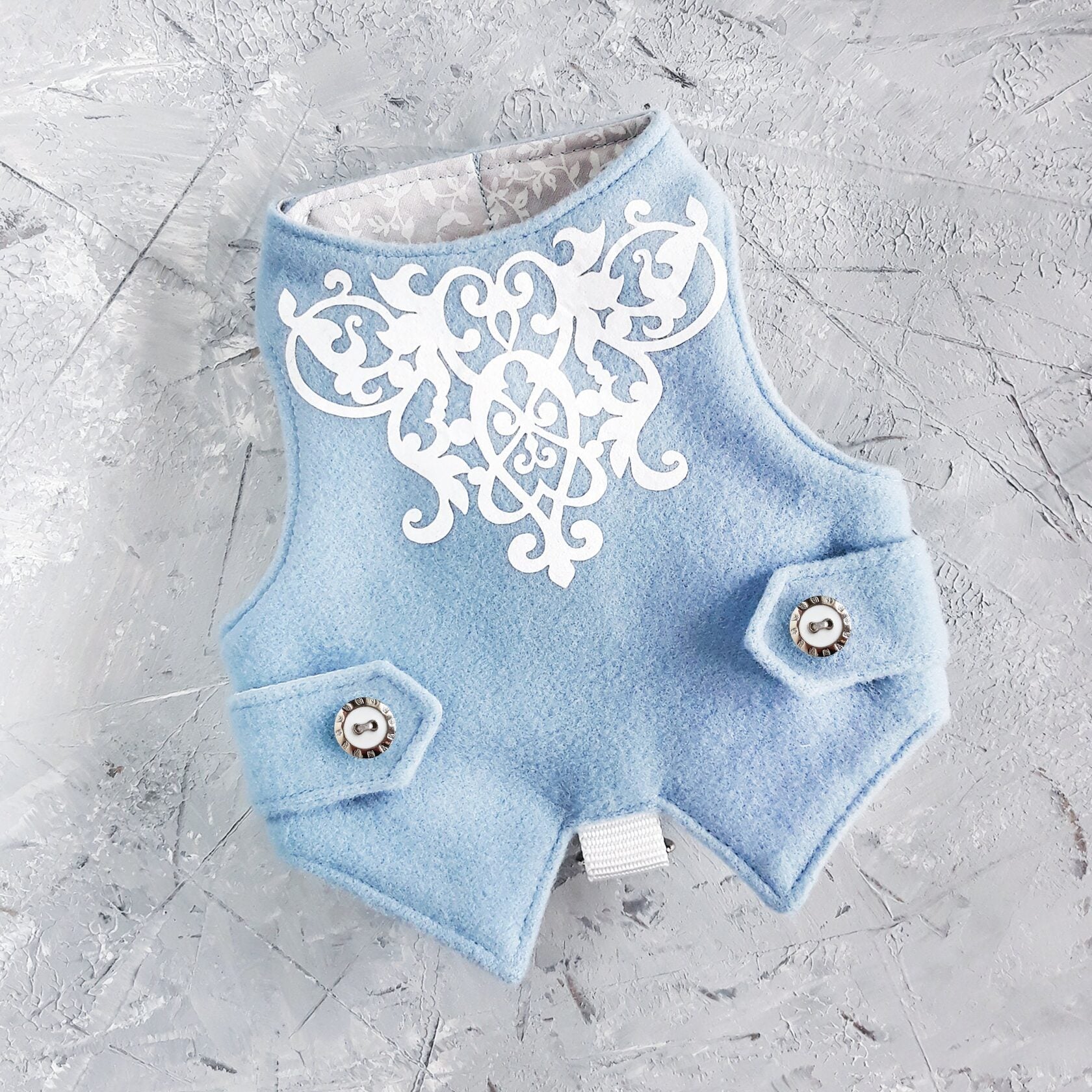 A soft blue cat harness, possibly wool, with a symmetrical white filigree design across the chest area. It includes small faux pocket flaps with decorative buttons and a silver-colored D-ring for leash attachment at the bottom. The interior lining is a light grey fabric with a subtle floral pattern.