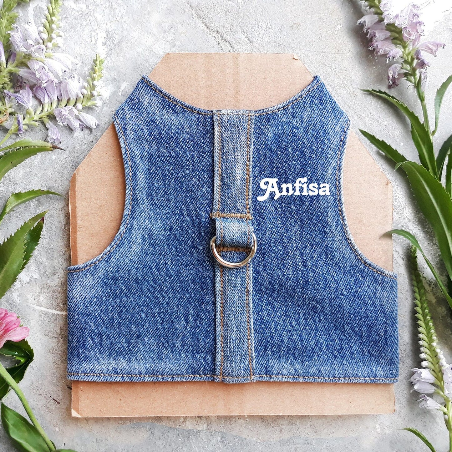 A cat harness made from blue denim fabric, featuring a central strap and a silver-colored D-ring with white cat`s name on it.