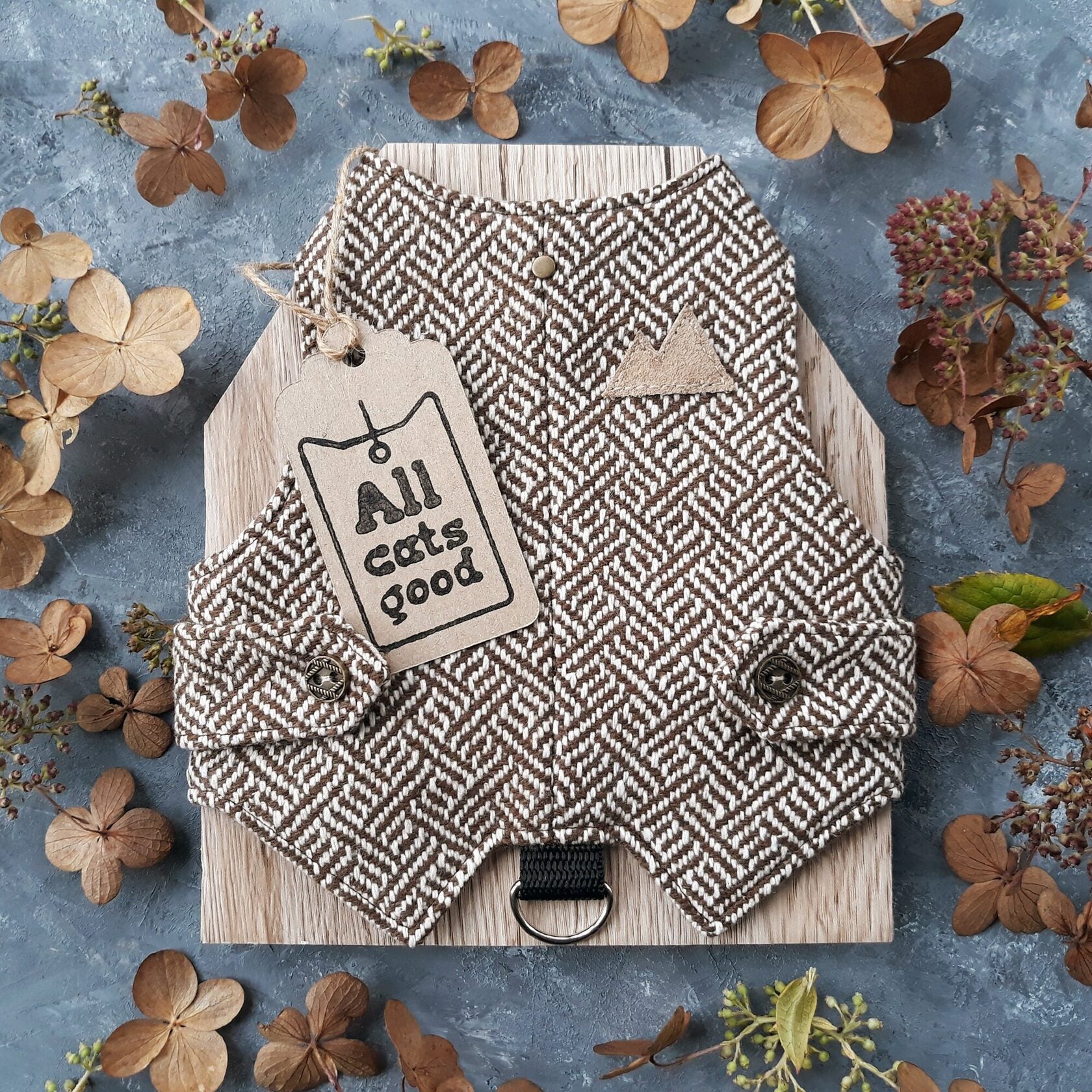 A cat harness made from brown tweed fabric with a distinctive geometric pattern. A functional D-ring connected by a black strap.
