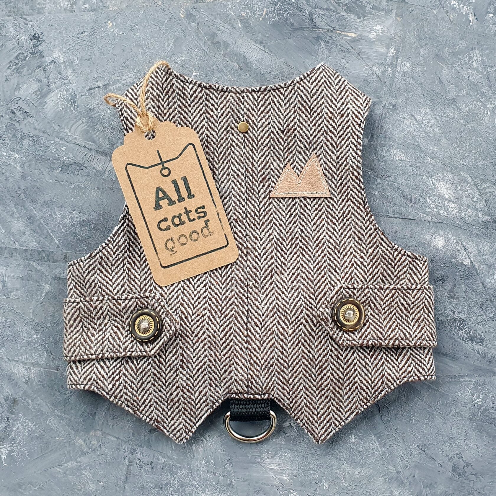 A brown and cream herringbone tweed cat harness with decorative bronze buttons, a brown faux leather mountain-shaped patch, and a black D-ring.