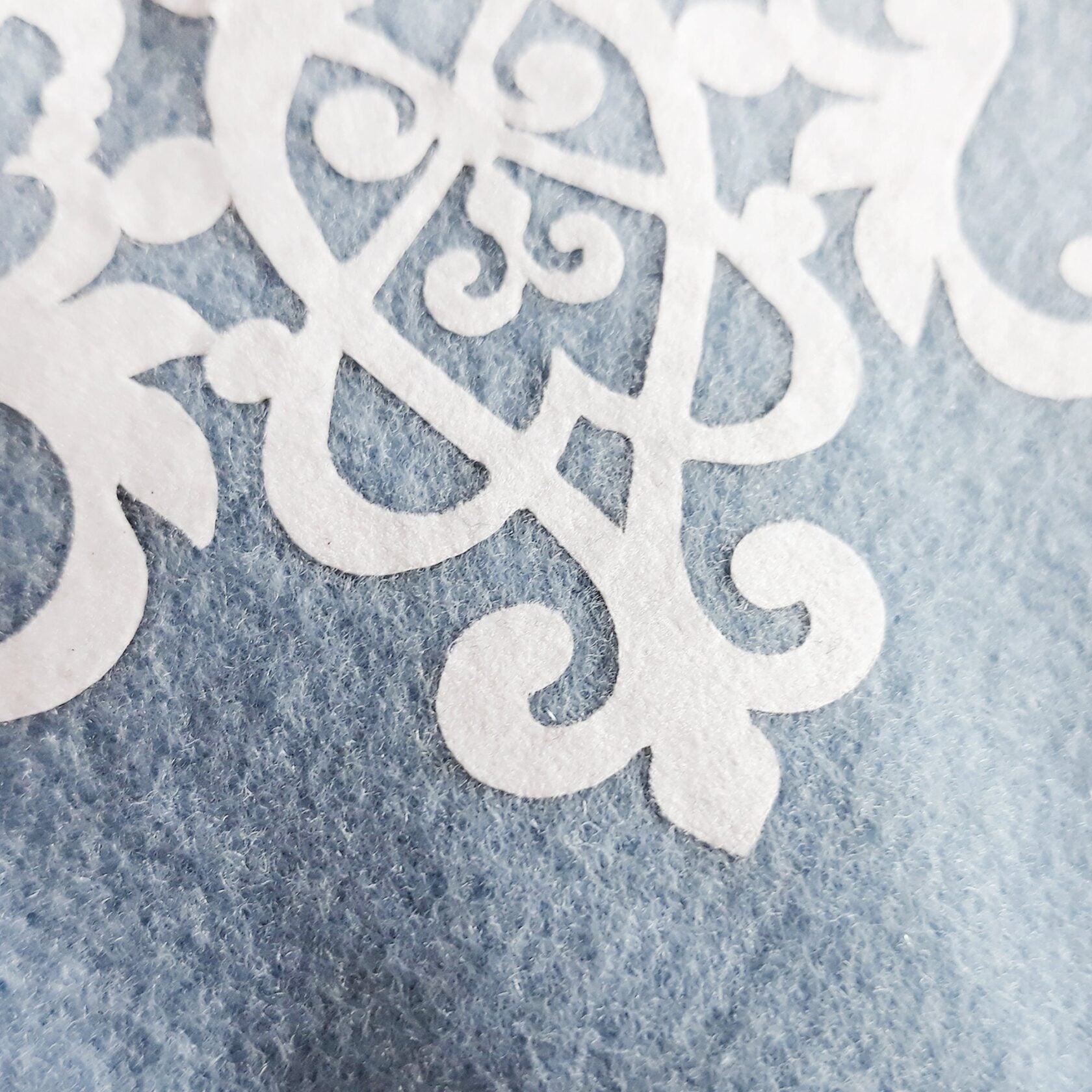Close-up of a white ornate swirl design on a light blue wool or felt cat harness.