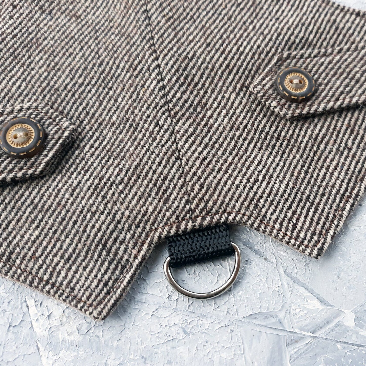 A detailed close-up shows decorative flap tabs with antique-style round buttons on a brown tweed cat harness with a diagonal stripes pattern. A silver metal D-ring attached to a black nylon strap is visible below.