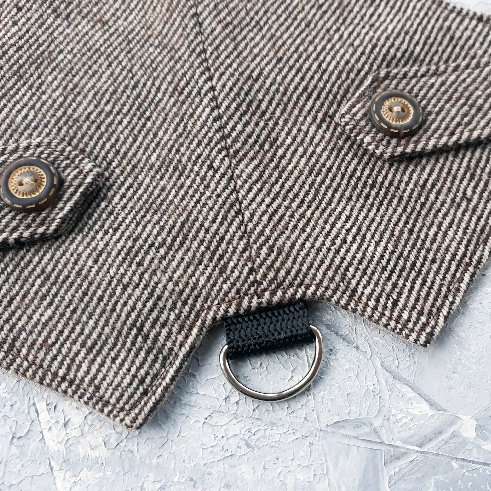 A detailed close-up shows decorative flap tabs with antique-style round buttons on a brown tweed cat harness with a diagonal stripes pattern. A silver metal D-ring attached to a black nylon strap is visible below.