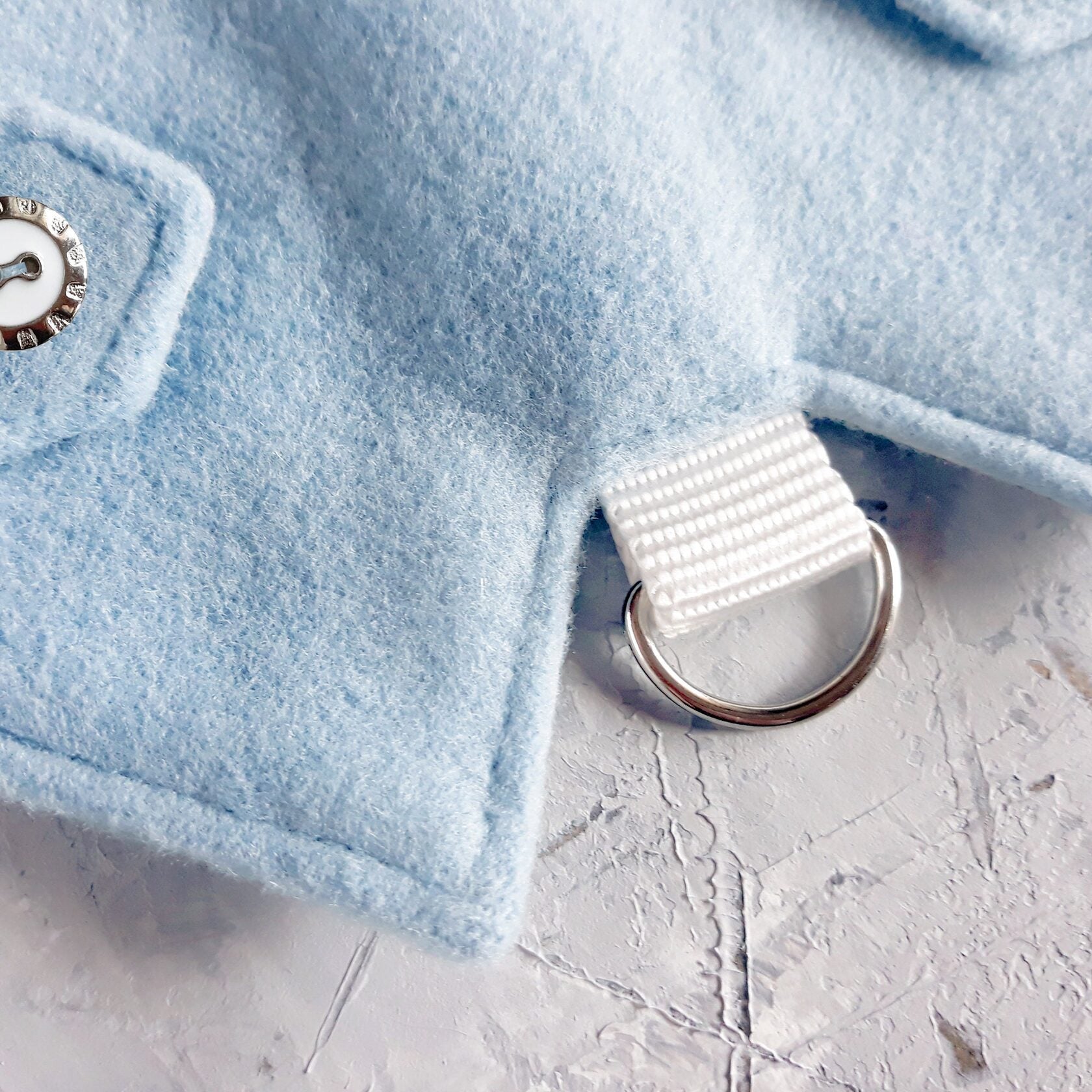Silver D-ring for leash attachment on a light blue cat harness, connected by a white fabric loop.
