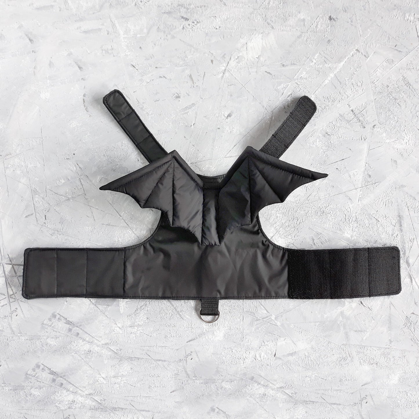 A black cat harness with a distinct design that includes bat wings extending from the back area. The harness has straps that would secure around the cat's neck and chest, fastened with Velcro.