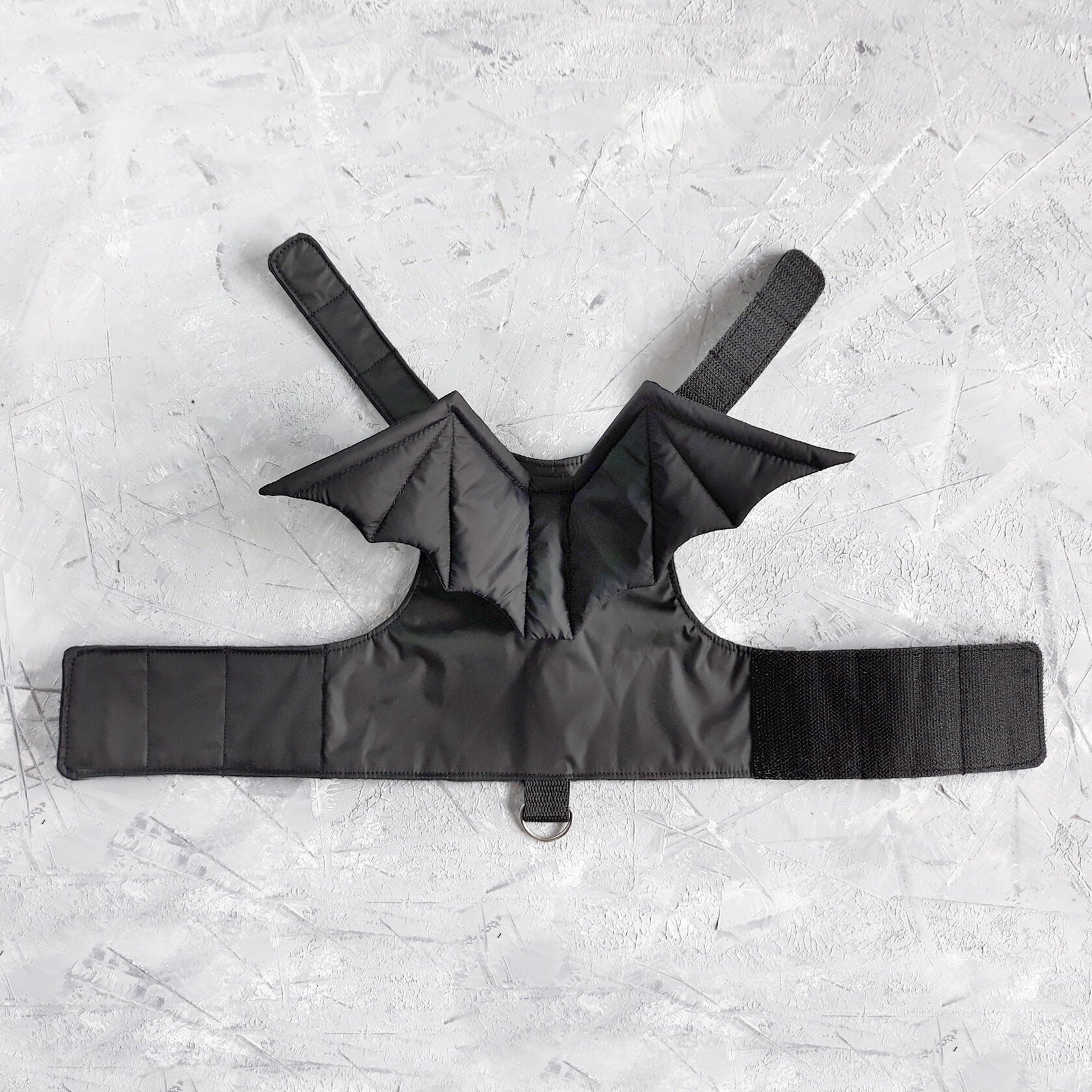 A black cat harness with a distinct design that includes bat wings extending from the back area. The harness has straps that would secure around the cat's neck and chest, fastened with Velcro.