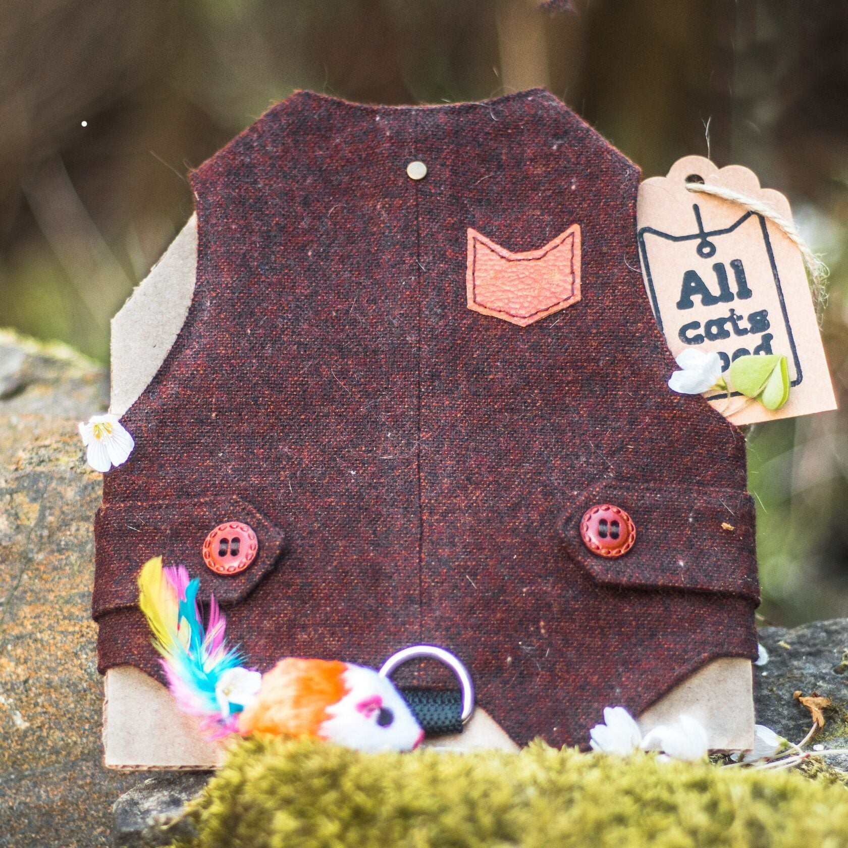 A burgundy tweed cat harness with a brown cat-shaped patch and decorative buttons, displayed on mossy rocks with a toy mouse and an "Allcatsgood" tag.