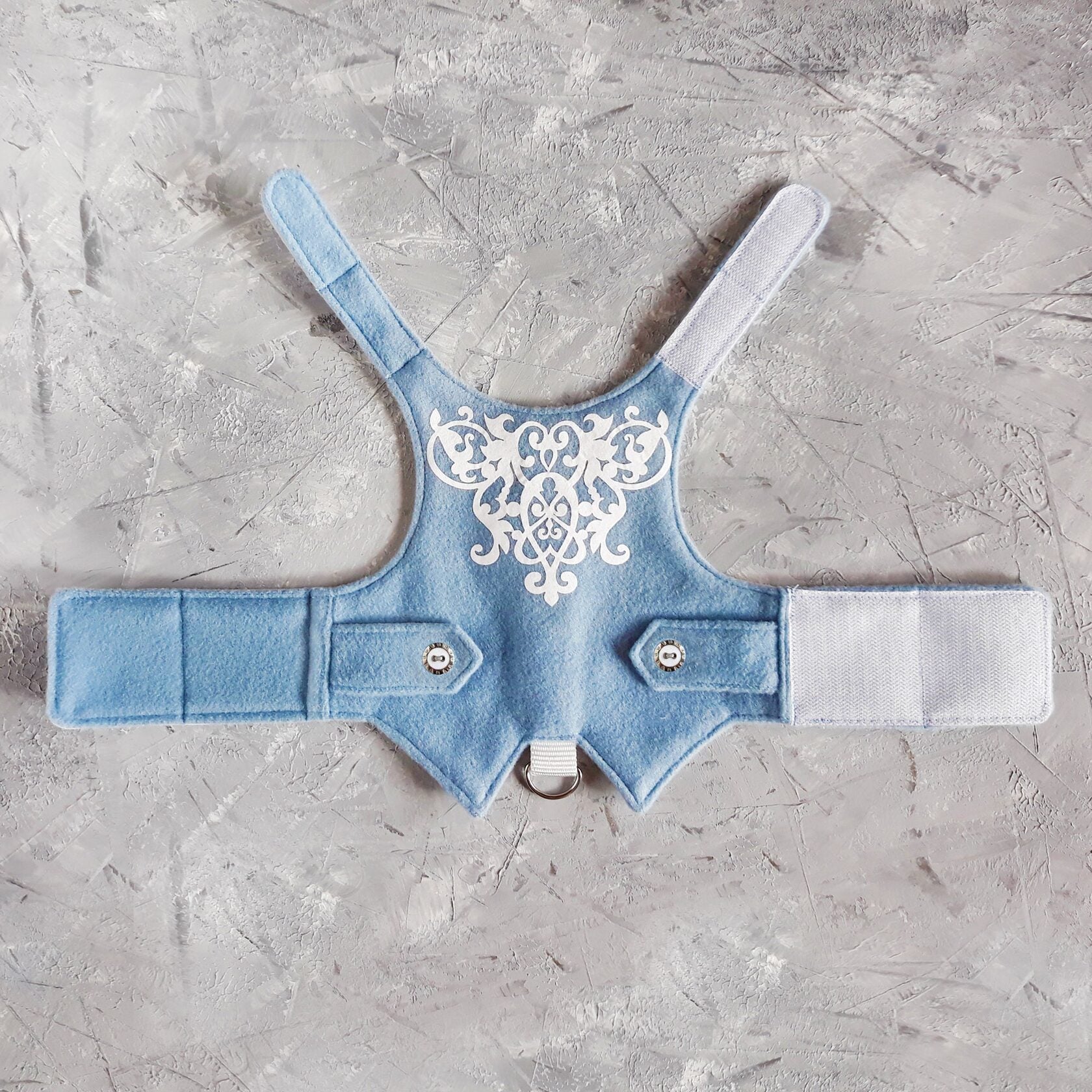 A soft blue cat harness, possibly wool, with a symmetrical white filigree design across the chest area. It includes small faux pocket flaps with decorative buttons and a silver-colored D-ring for leash attachment at the bottom. The interior lining is a light grey fabric with a subtle floral pattern.