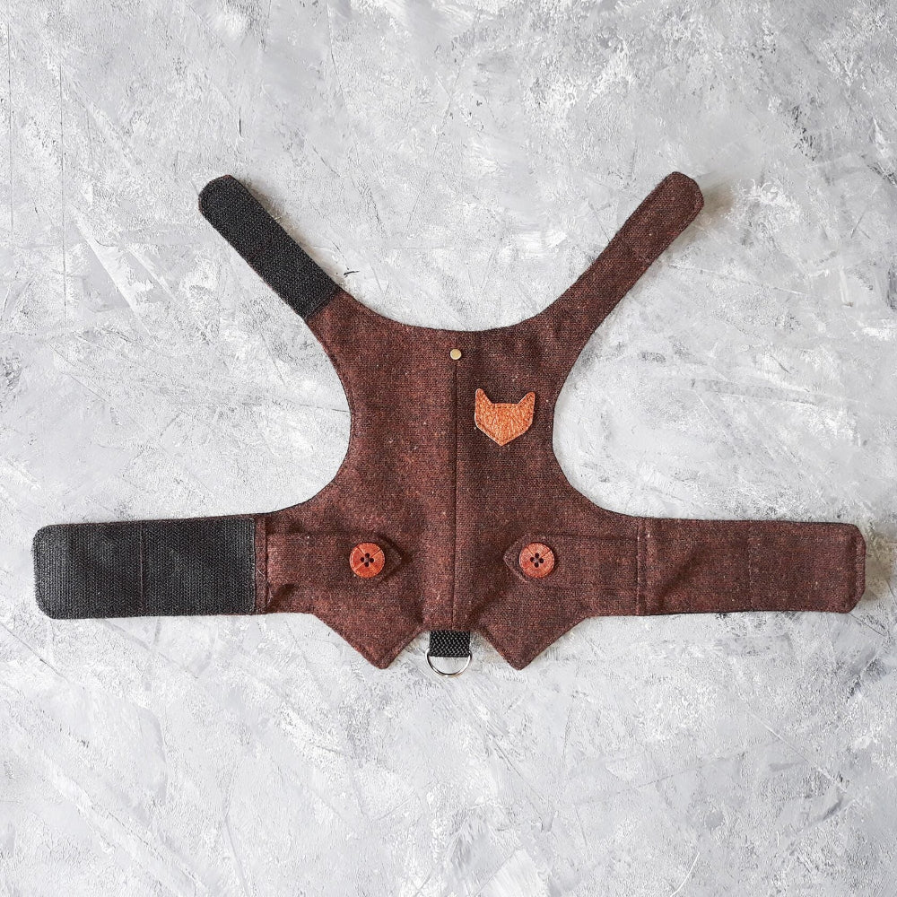 A flat lay of a burgundy tweed cat harness with a brown cat-shaped patch, decorative buttons, black straps, and a D-ring.