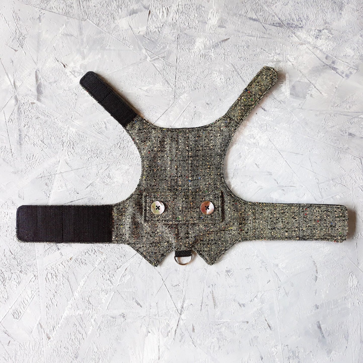 A flat lay of a grey tweed cat harness with colorful flecks, decorative mother-of-pearl buttons, black Velcro straps, and a silver D-ring.