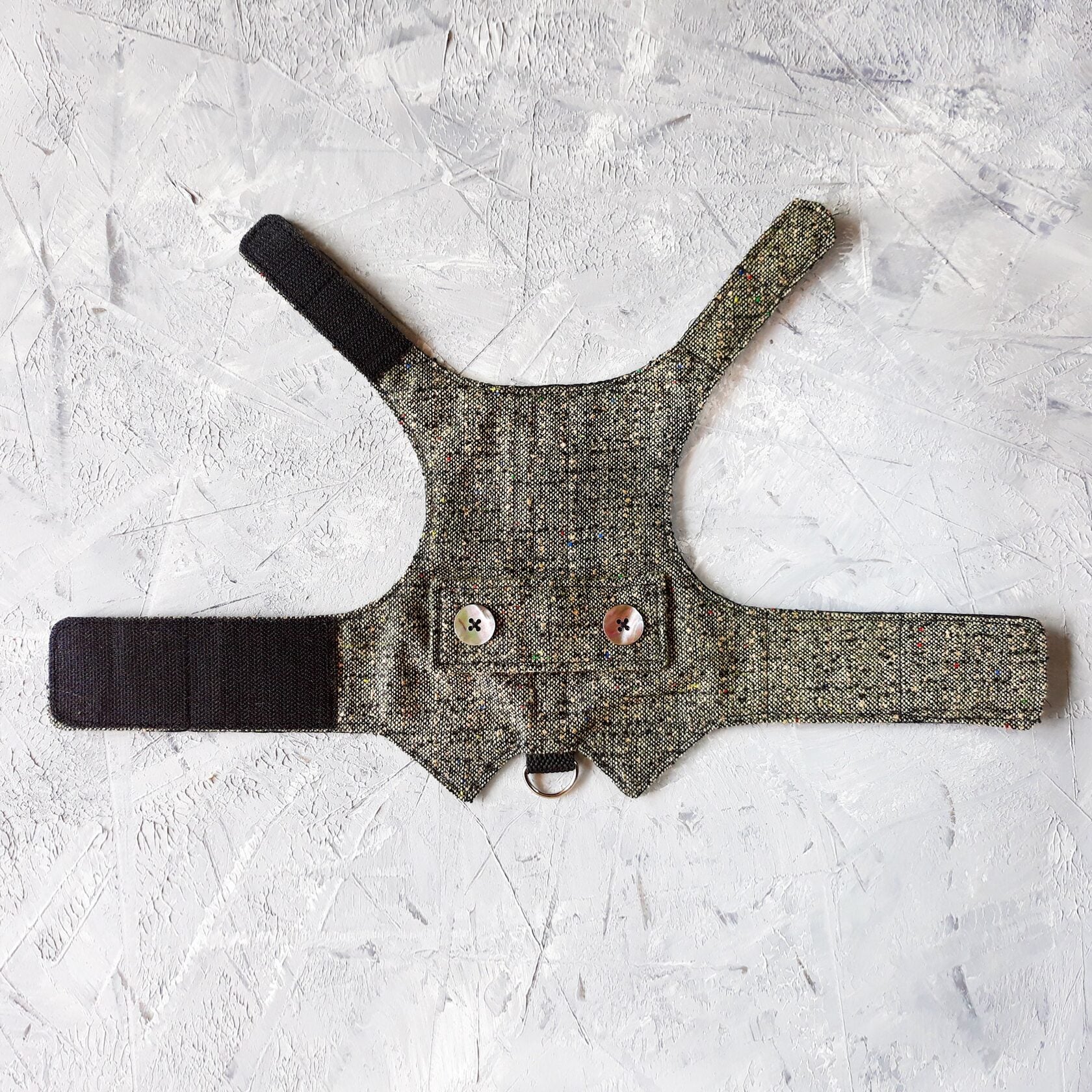 A flat lay of a grey tweed cat harness with colorful flecks, decorative mother-of-pearl buttons, black Velcro straps, and a silver D-ring.