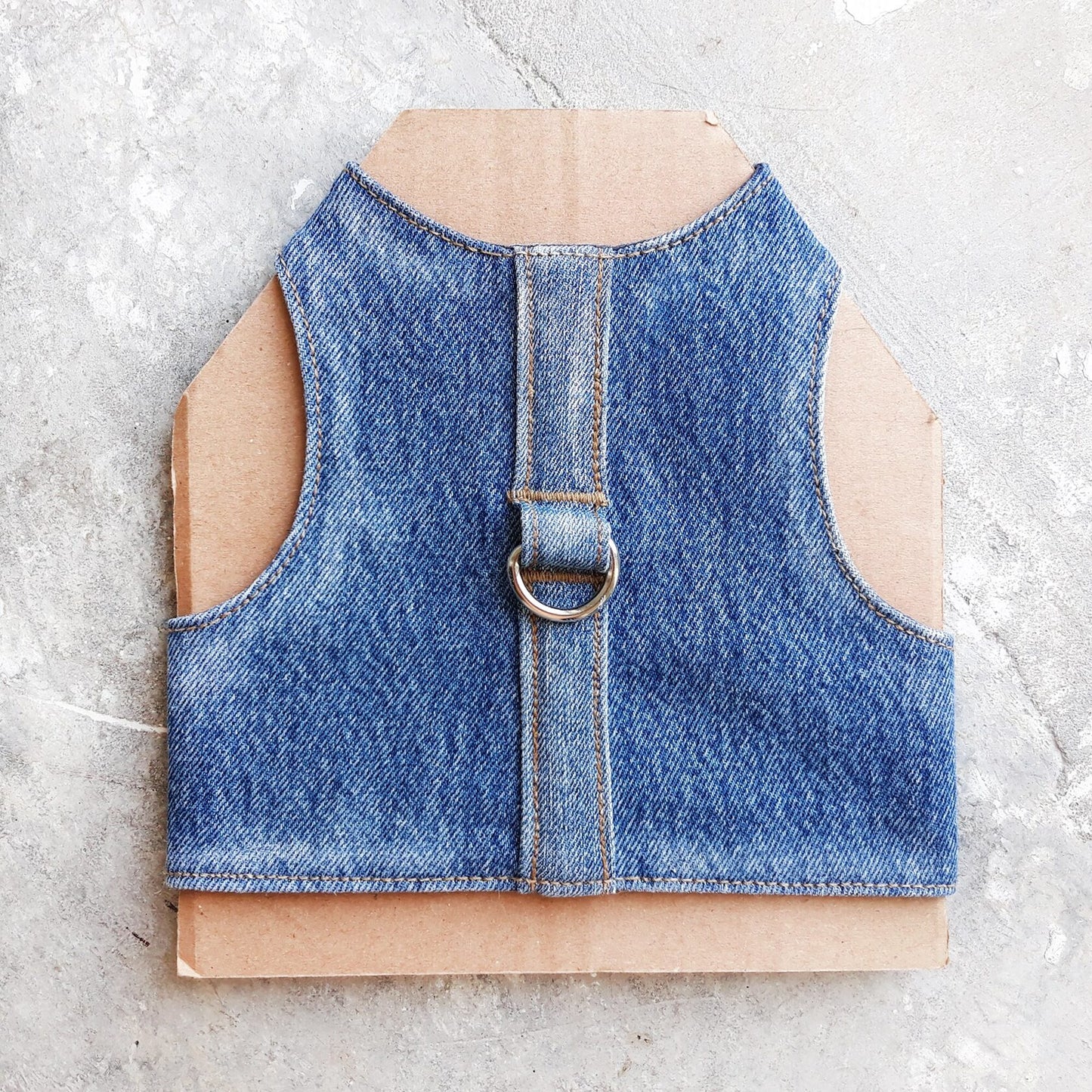 A cat harness made from blue denim fabric, featuring a central strap and a silver-colored D-ring.