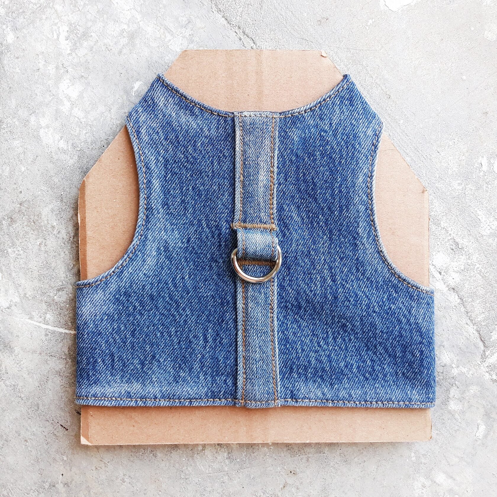 A cat harness made from blue denim fabric, featuring a central strap and a silver-colored D-ring.