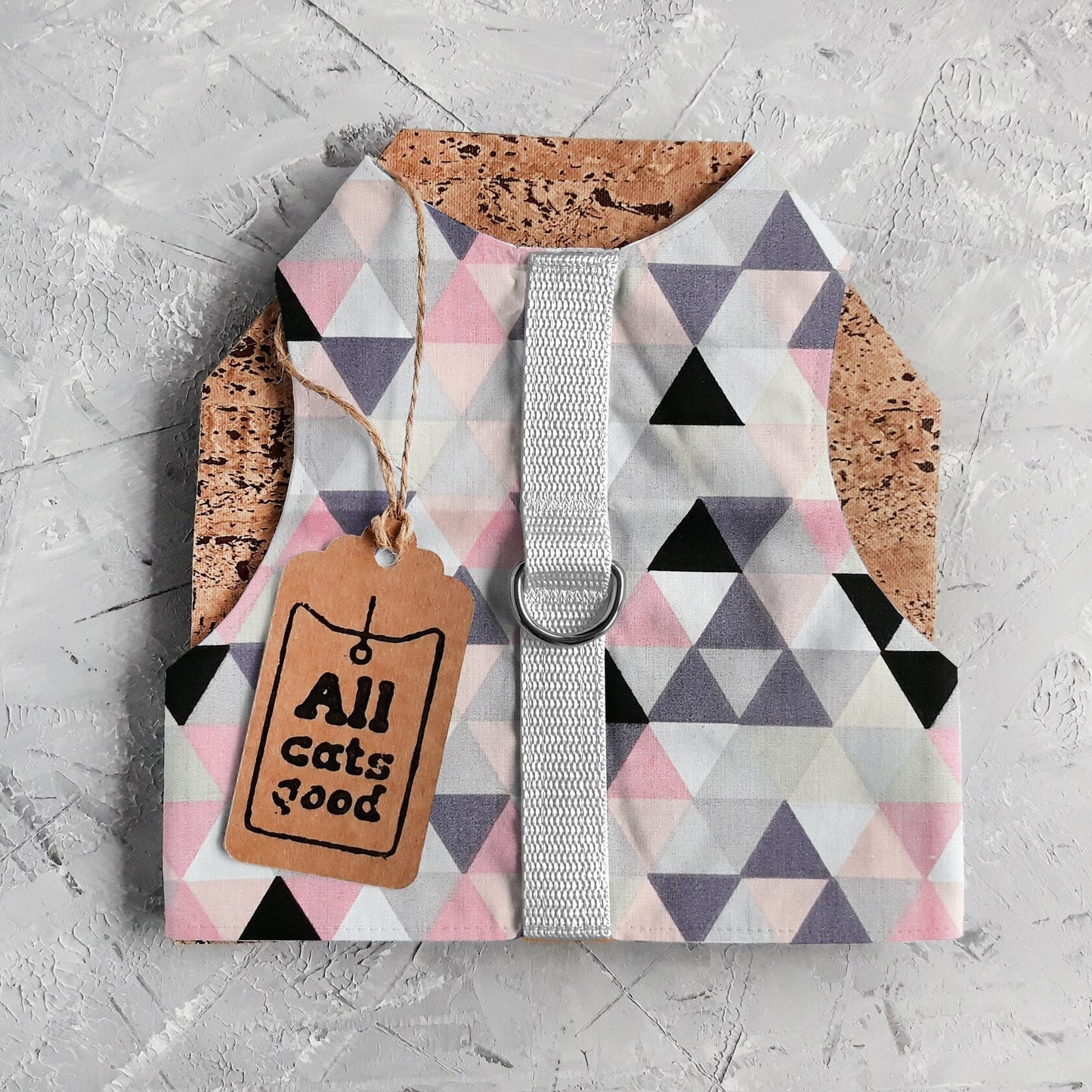 A cat harness with a geometric triangle pattern in pink, grey, white, and black, a silver D-ring, a light grey vertical strap, cork side panels, and an Allcatsgood tag.