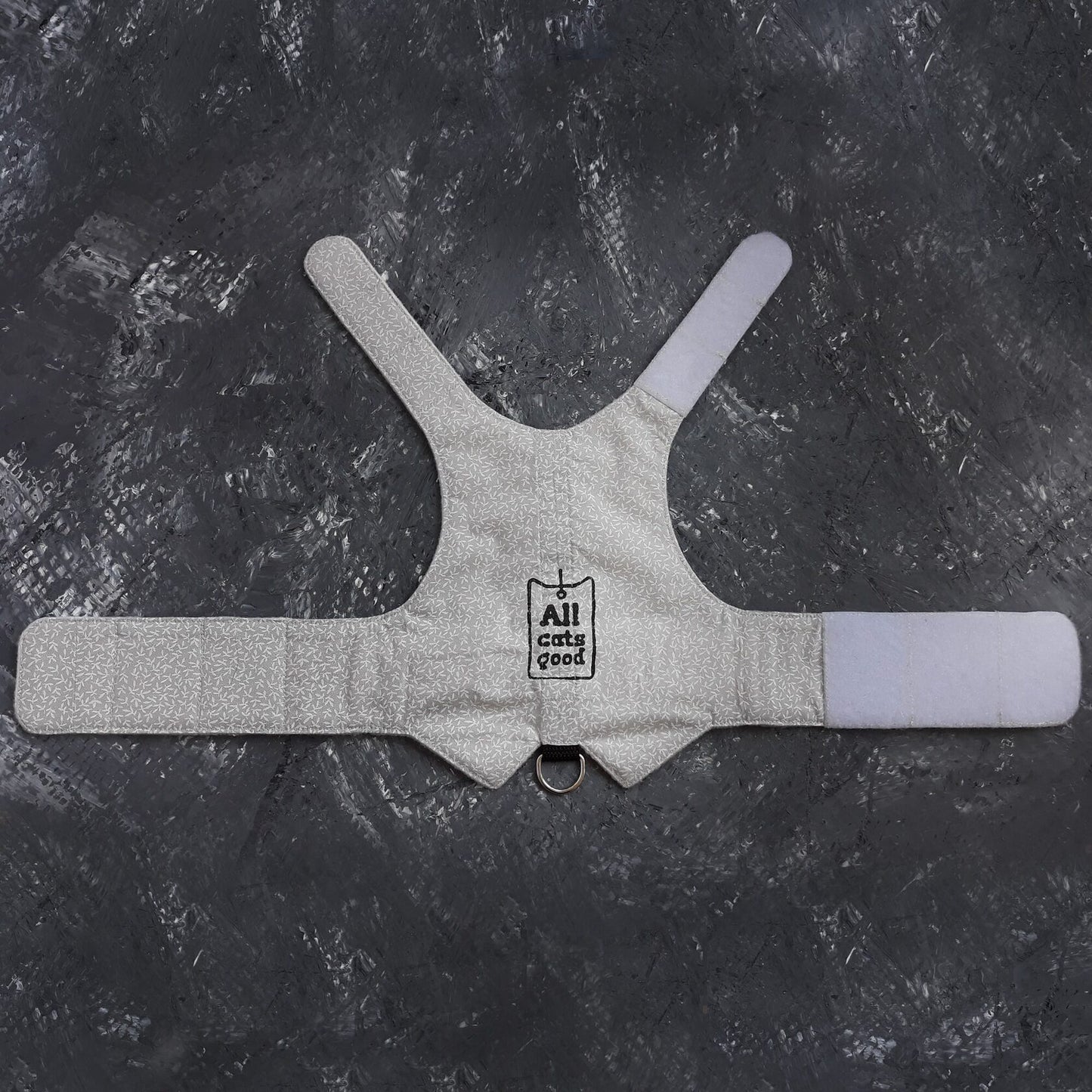 A white cat harness, described as is laid out to display its vest shape, white Velcro straps for adjustability, snap closures for fastening, and a metal D-ring for attaching a leash.