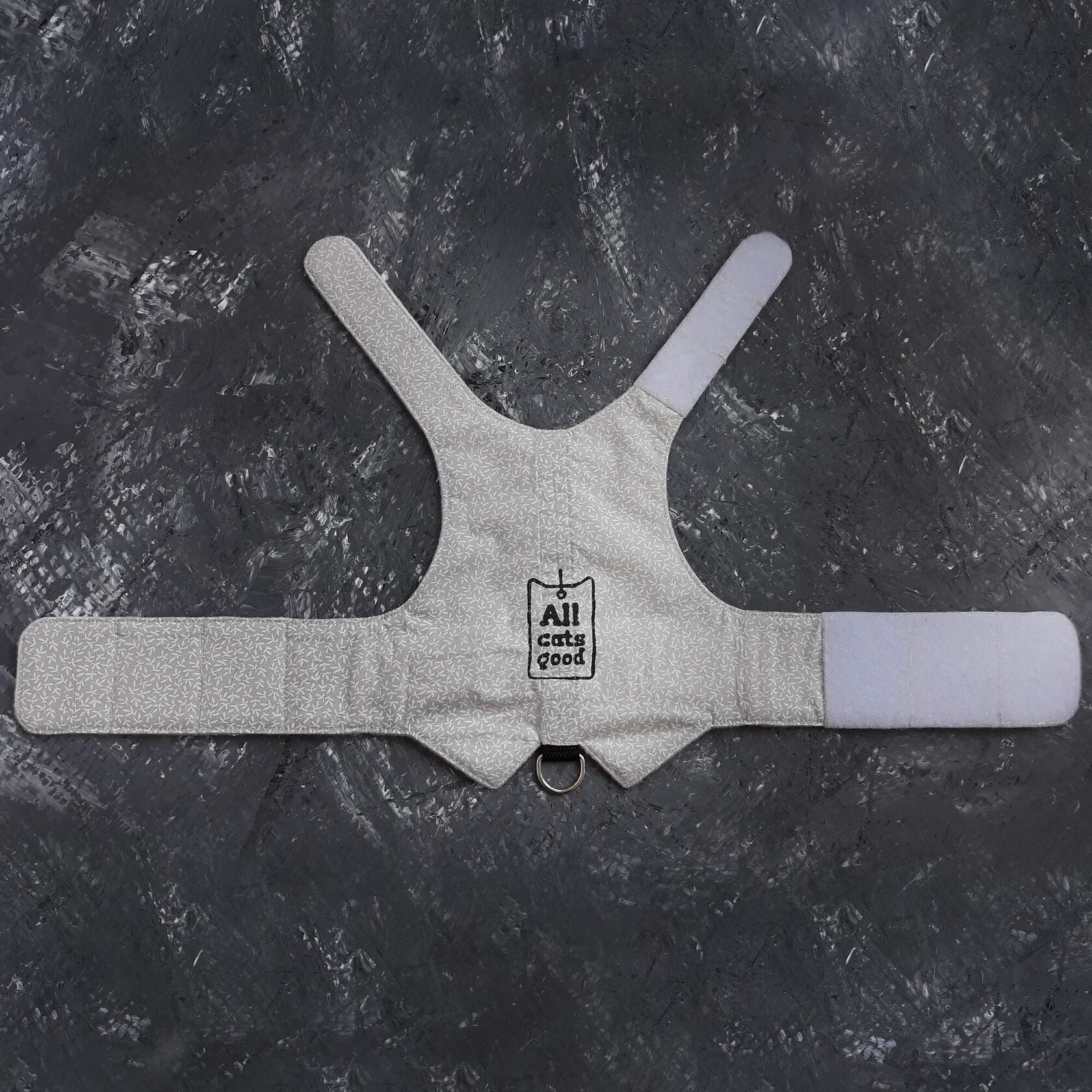 A white cat harness, described as is laid out to display its vest shape, white Velcro straps for adjustability, snap closures for fastening, and a metal D-ring for attaching a leash.