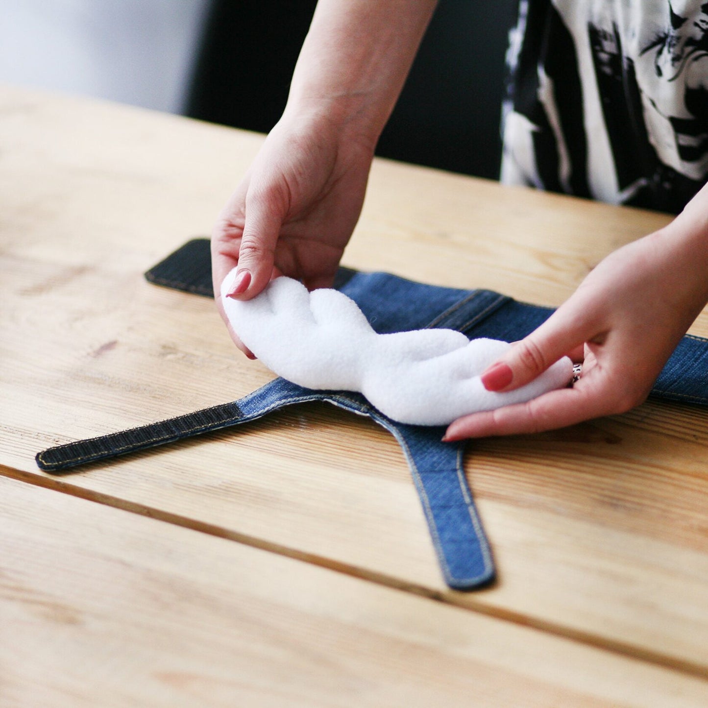 Hands attaching removable white angel wings to a blue upcycled denim cat harness on a wooden table.