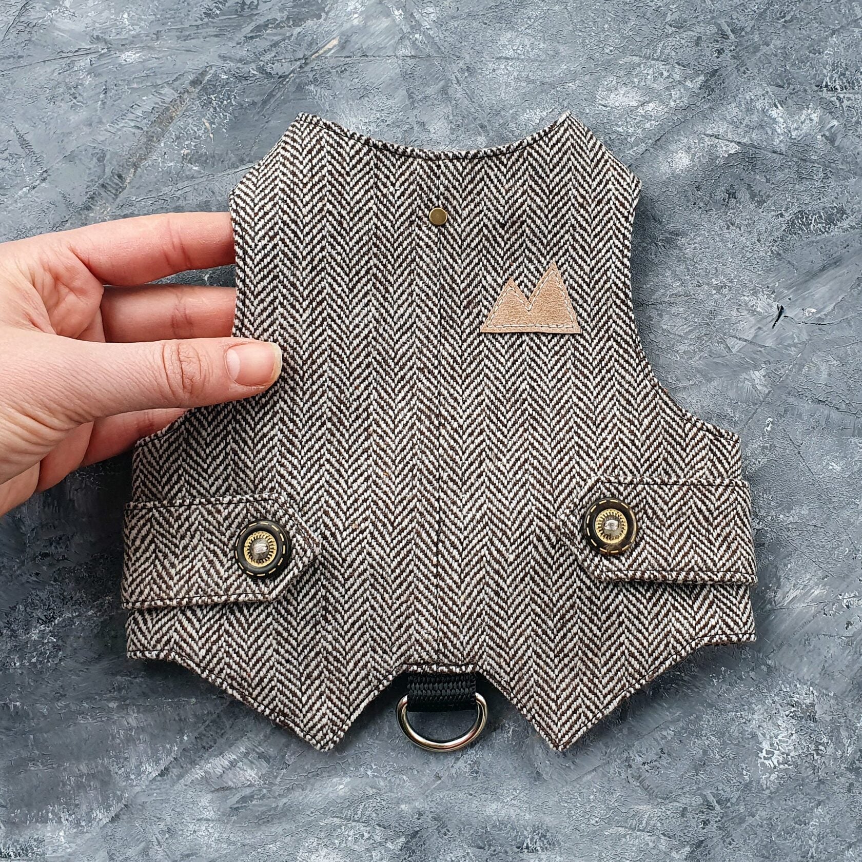 A brown and cream herringbone tweed cat harness with decorative bronze buttons, a brown faux leather mountain-shaped patch, and a black D-ring.